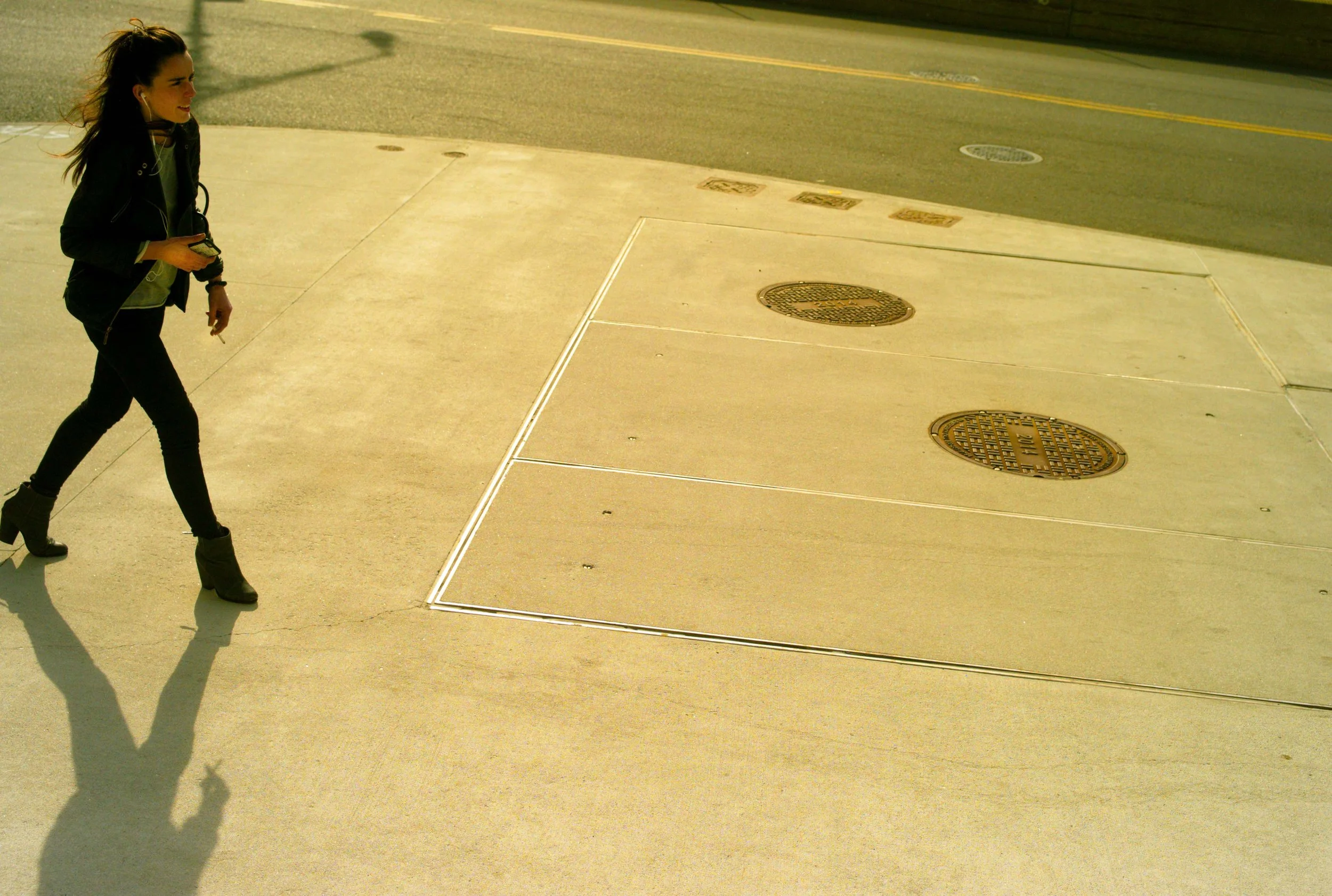 A young woman with dark hair in a ponytail, wearing black pants and black boots, is walking on a sidewalk while looking to her left. She is holding a phone and smiling. The sidewalk has a white outline around a section with round metal utility covers