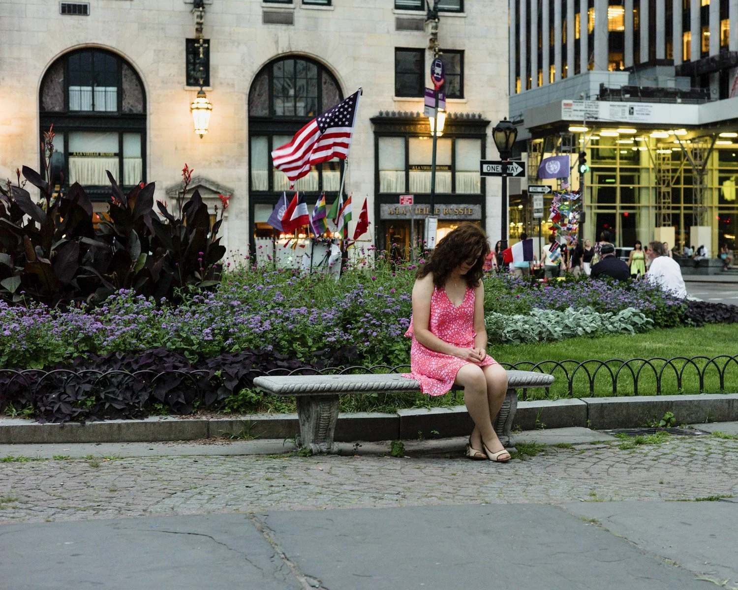 A woman in a pink dress sitting on a park bench surrounded by purple flowers and plants, with city buildings and flags in the background.
