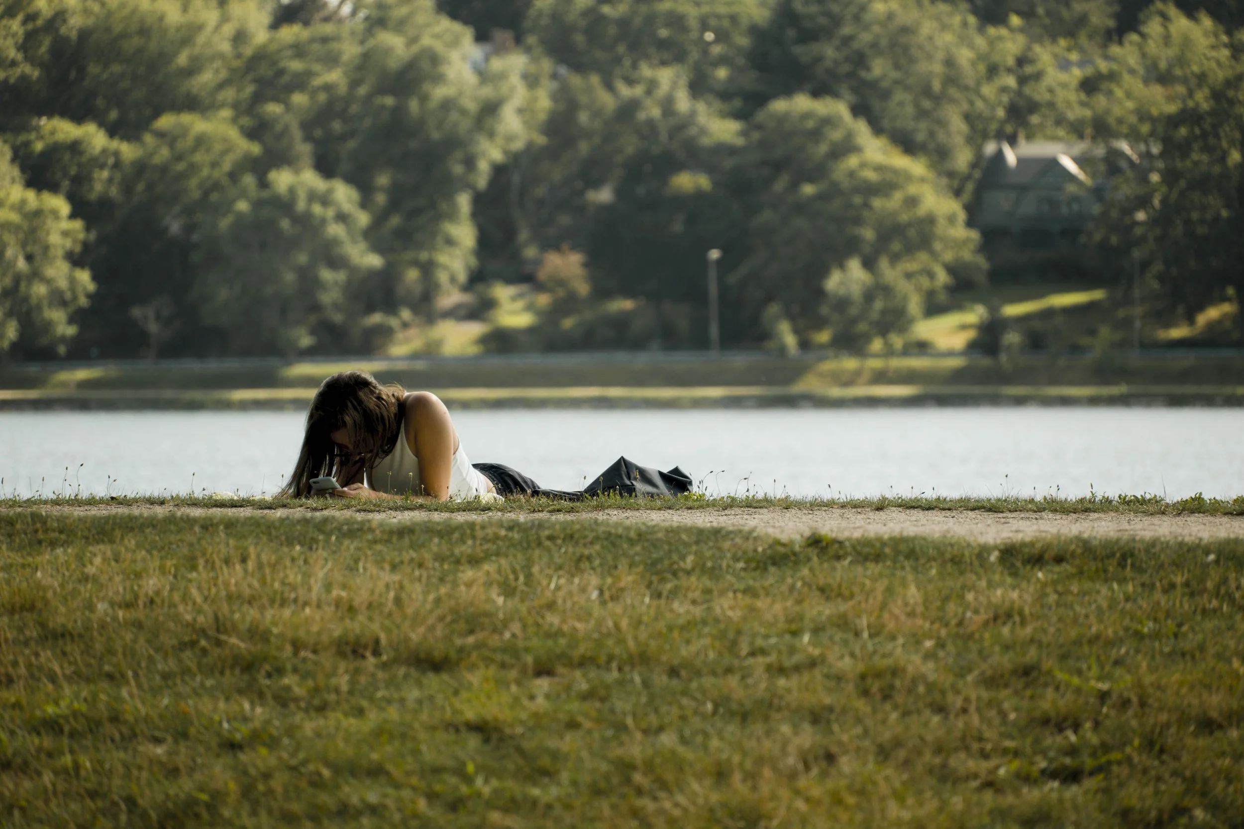 A woman lying on the grass by a lake, looking at her phone, with trees and houses in the background.