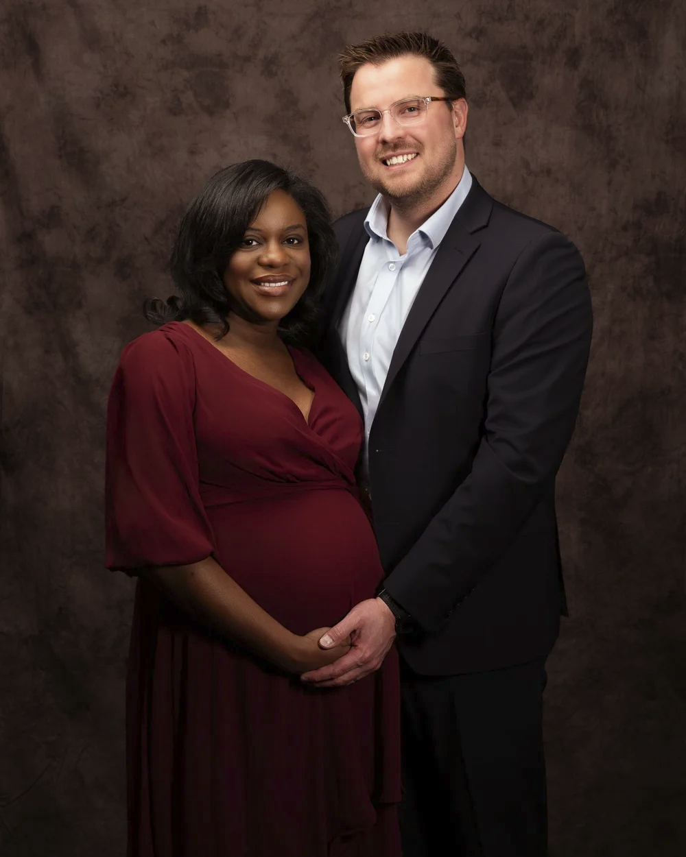 A pregnant woman in a red dress and a man in a black suit pose together, smiling, with the woman's hand on her baby bump against a brown textured background.