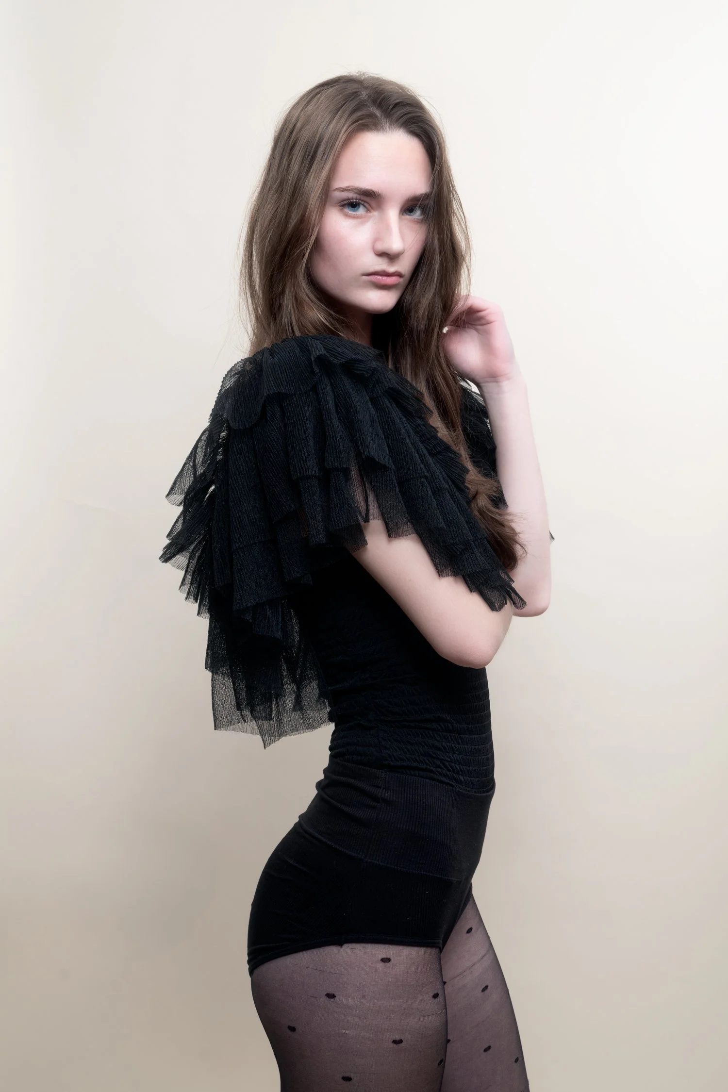 Young woman with long brown hair wearing a black, ruffled, tulle top, black fitted shorts, patterned sheer tights, and posing against a plain light background.
