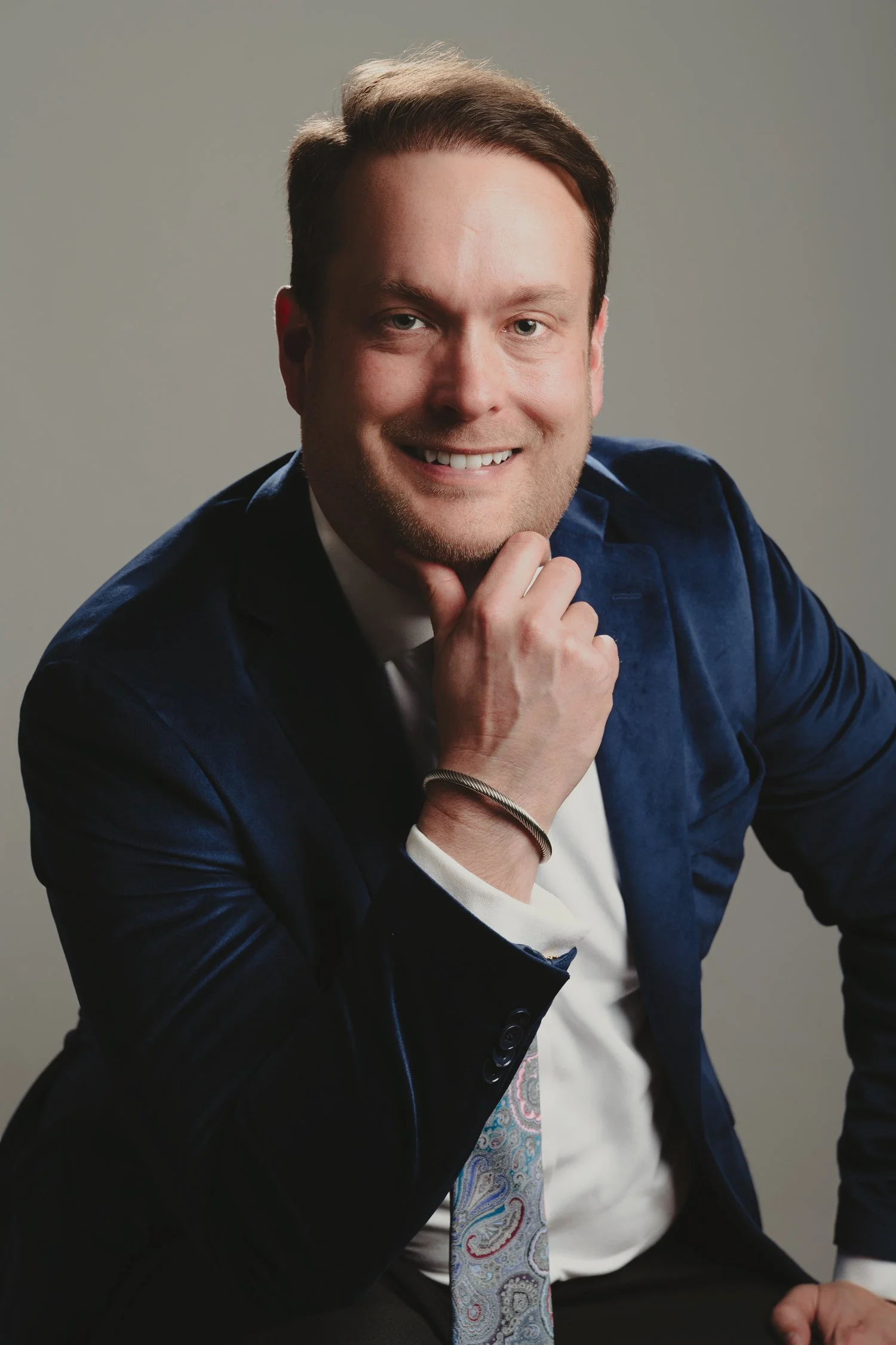 A man in a navy blue suit and colorful tie smiling with his hand resting on his chin, posed against a plain gray background.