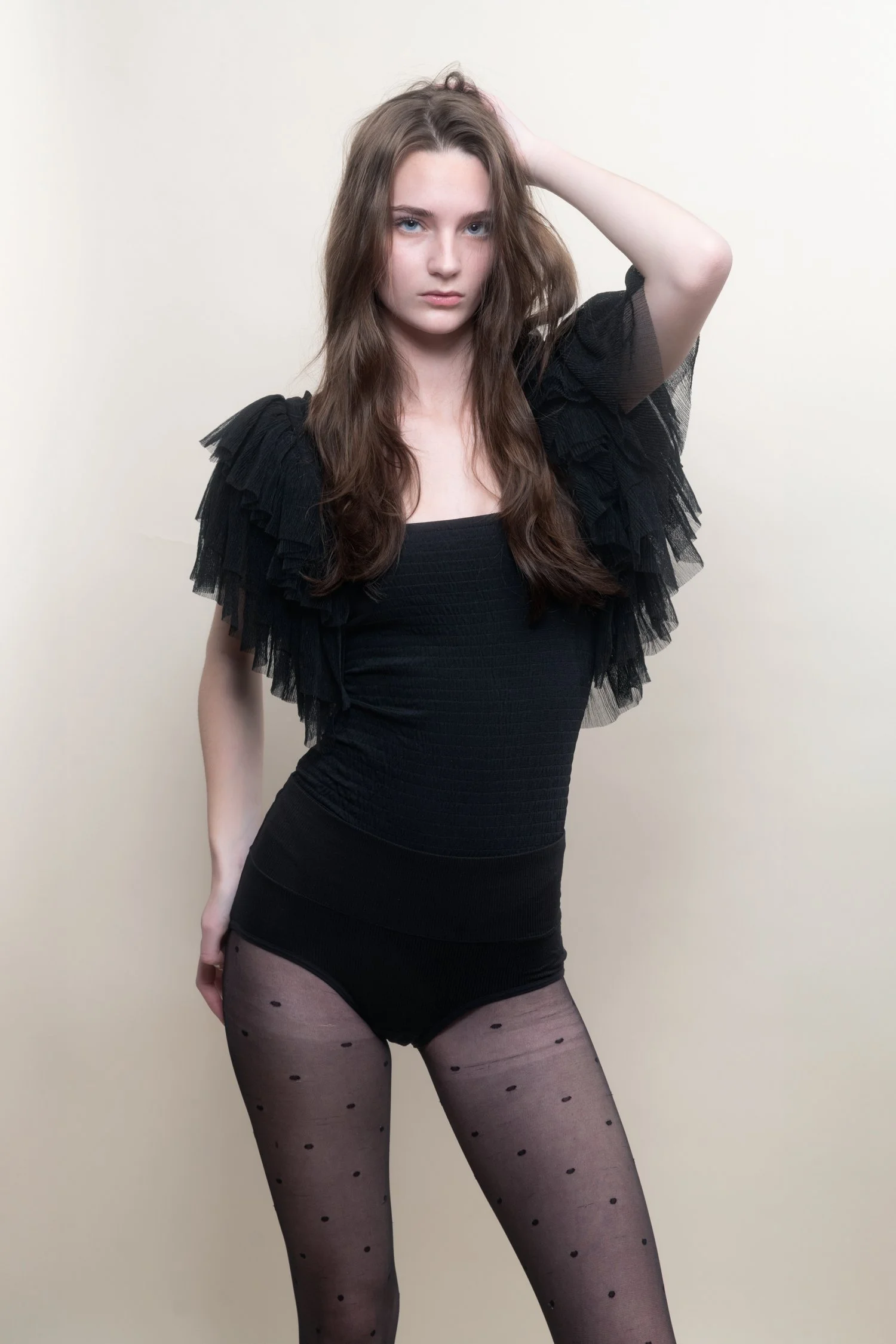Young woman with long brown hair wearing a black ruffled top, sheer polka dot tights, and posing with one hand in her hair against a plain neutral background.