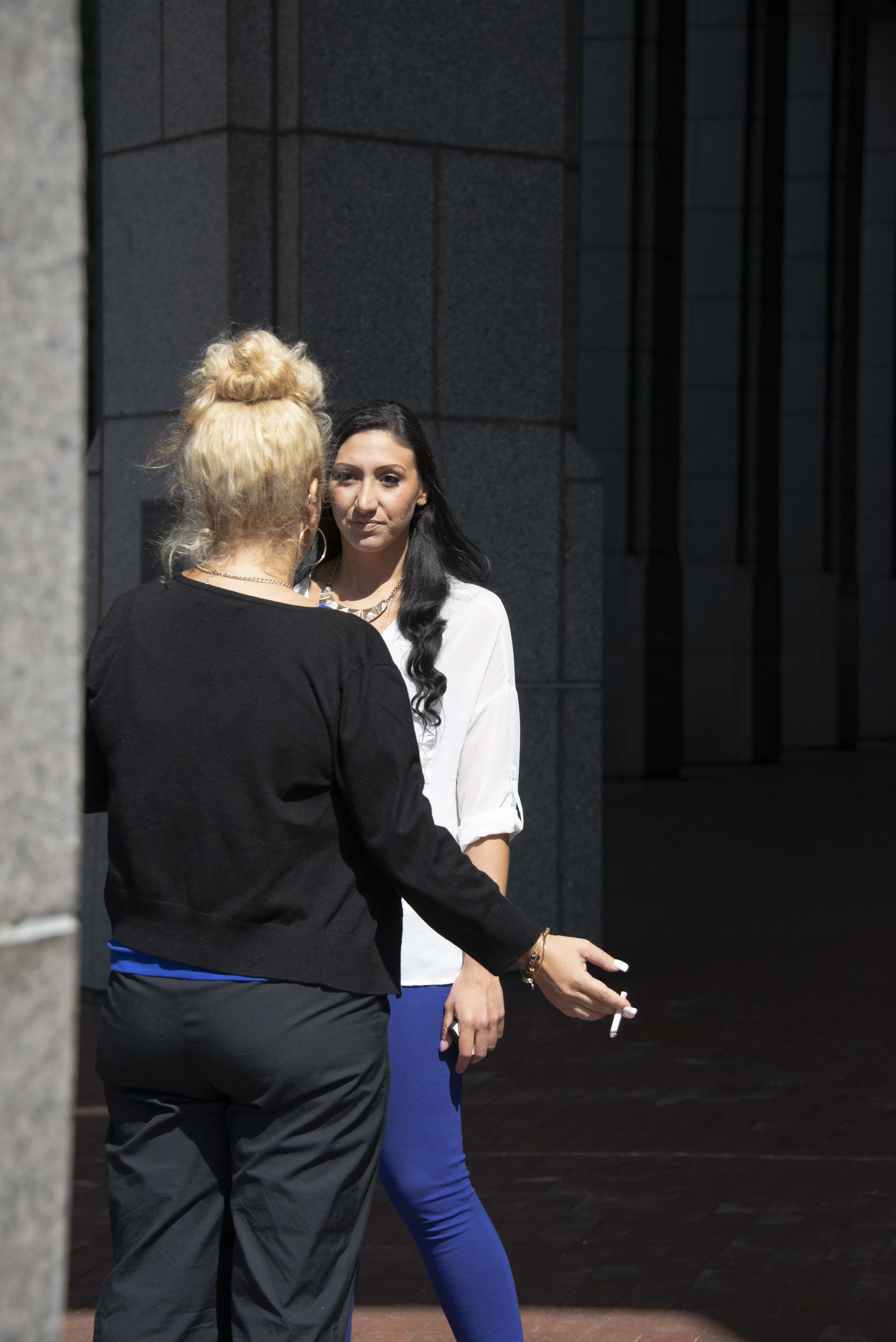 Two women talking outdoors near a building with dark gray walls. The woman facing away has blonde hair in a bun and is wearing a black top. The woman facing forward has long black hair, wears a white top, and blue pants, and holds a cigarette in her 