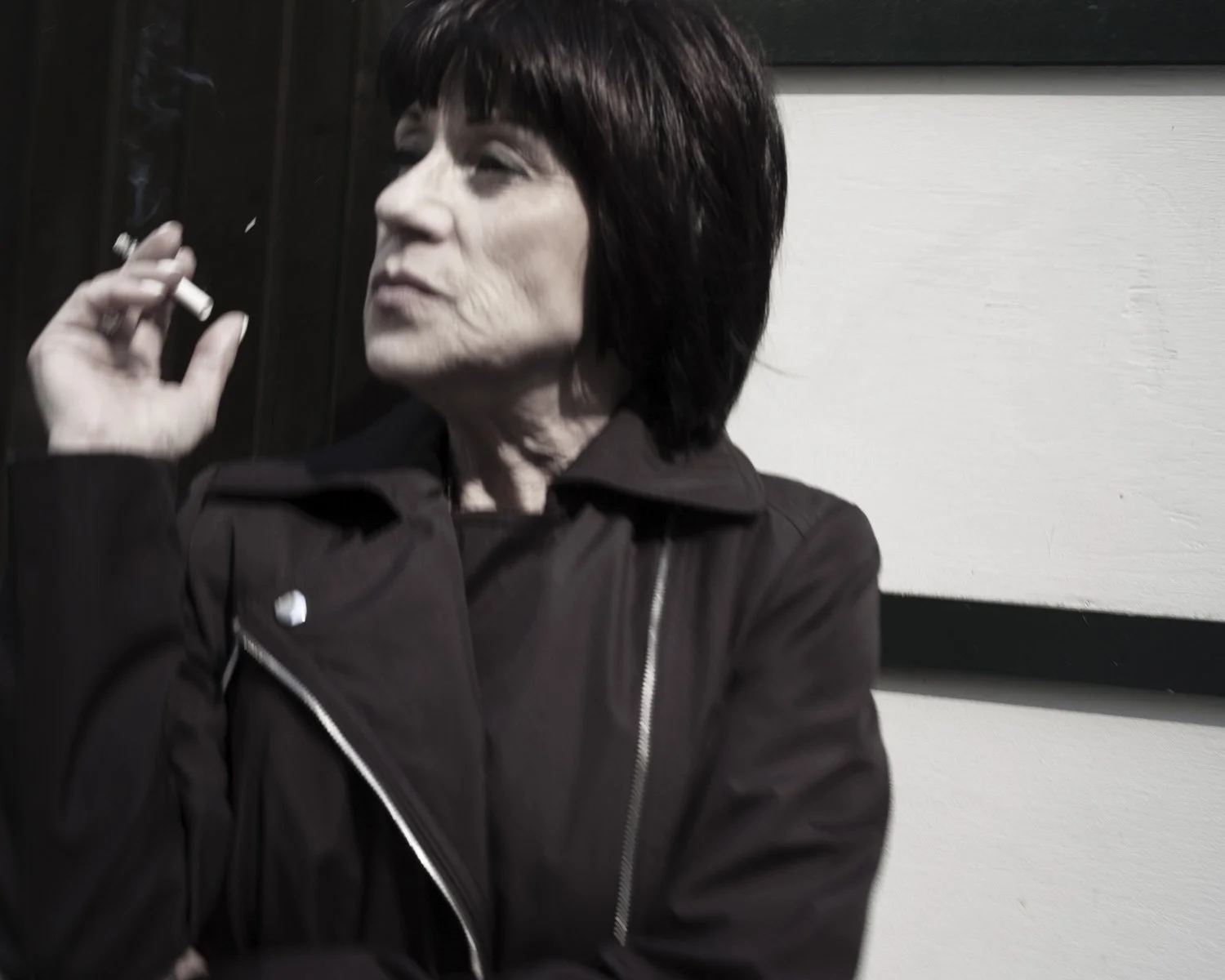 A middle-aged woman with dark hair and light skin, wearing a black jacket, smoking a cigarette. She is sitting against a black and white background with wood paneling.