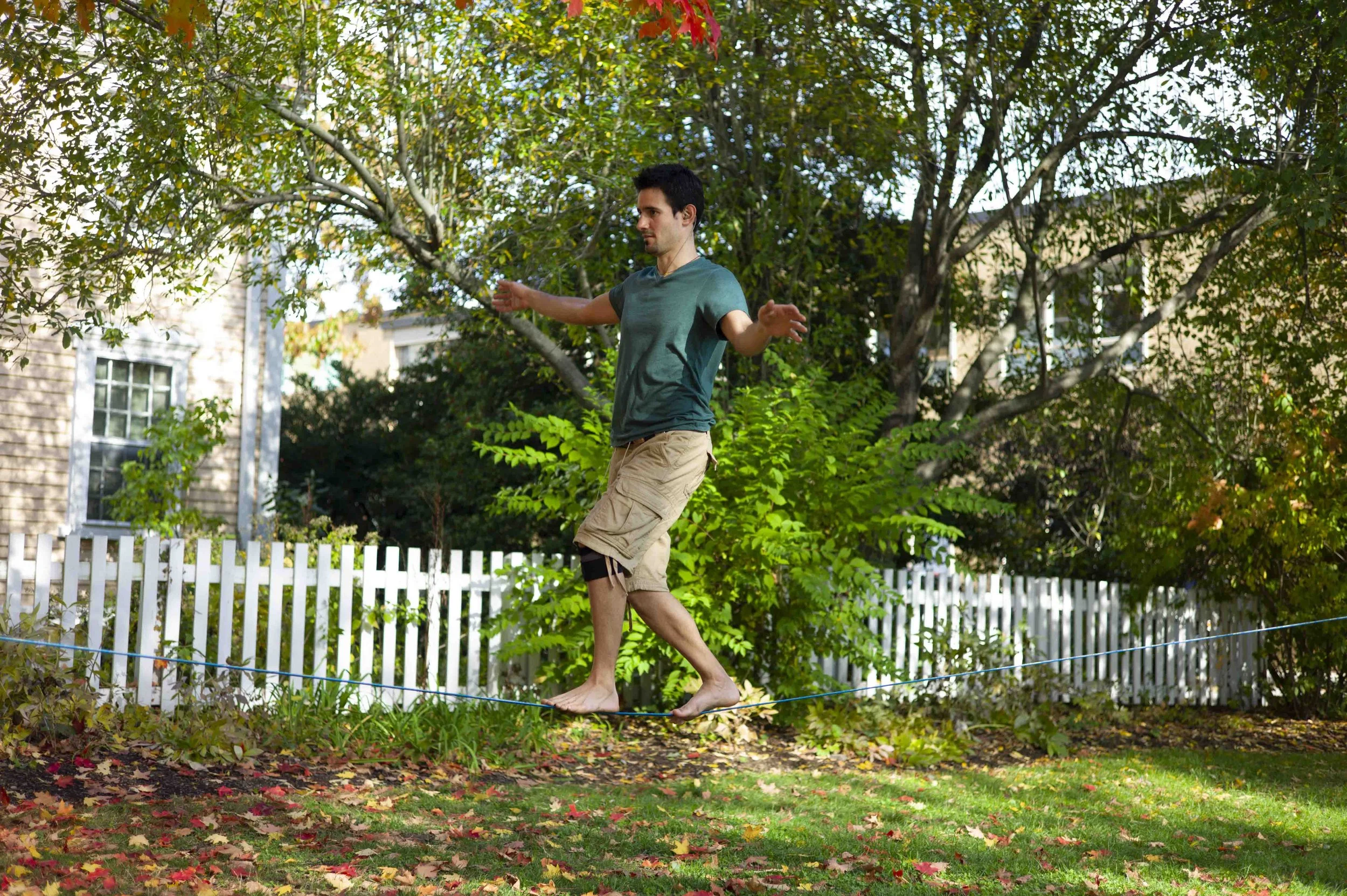 Man walking barefoot on a slackline in a backyard, surrounded by trees and fall foliage.