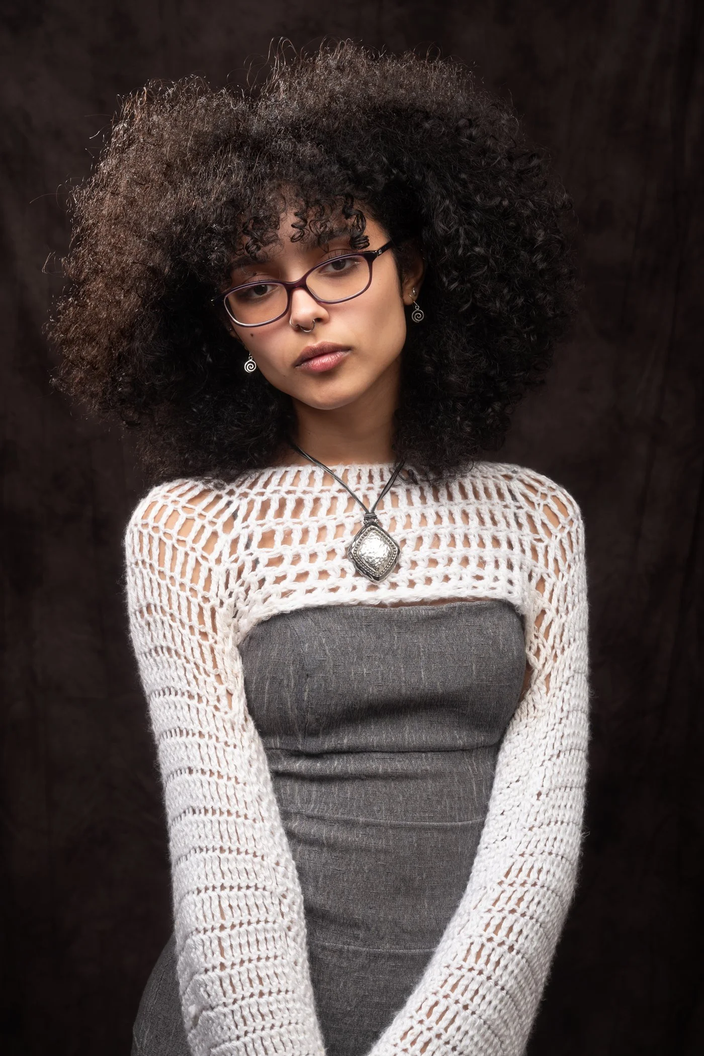 Portrait of  young ethnic woman with black, tightly-curled hair and heavy-rimmed glasses. She’s seen against a dark backgrdrop, wearing a white-knit dress with a gray lower portion.