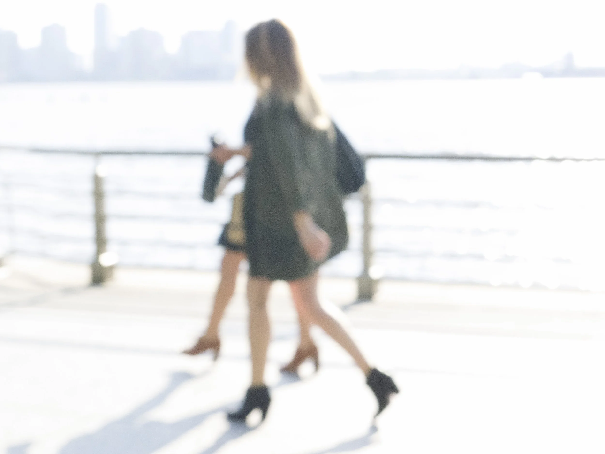 Blurry image of a woman with long hair, wearing a dark jacket and shorts, standing near a body of water with a city skyline in the background.