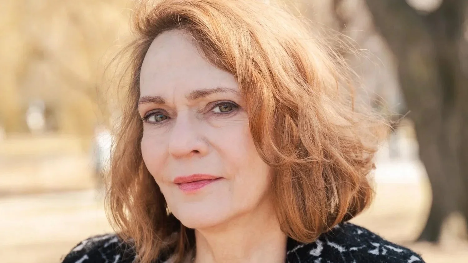 Closeup shot of a woman with auburn-color hair in a park setting, makes eye-contact with the camera.
