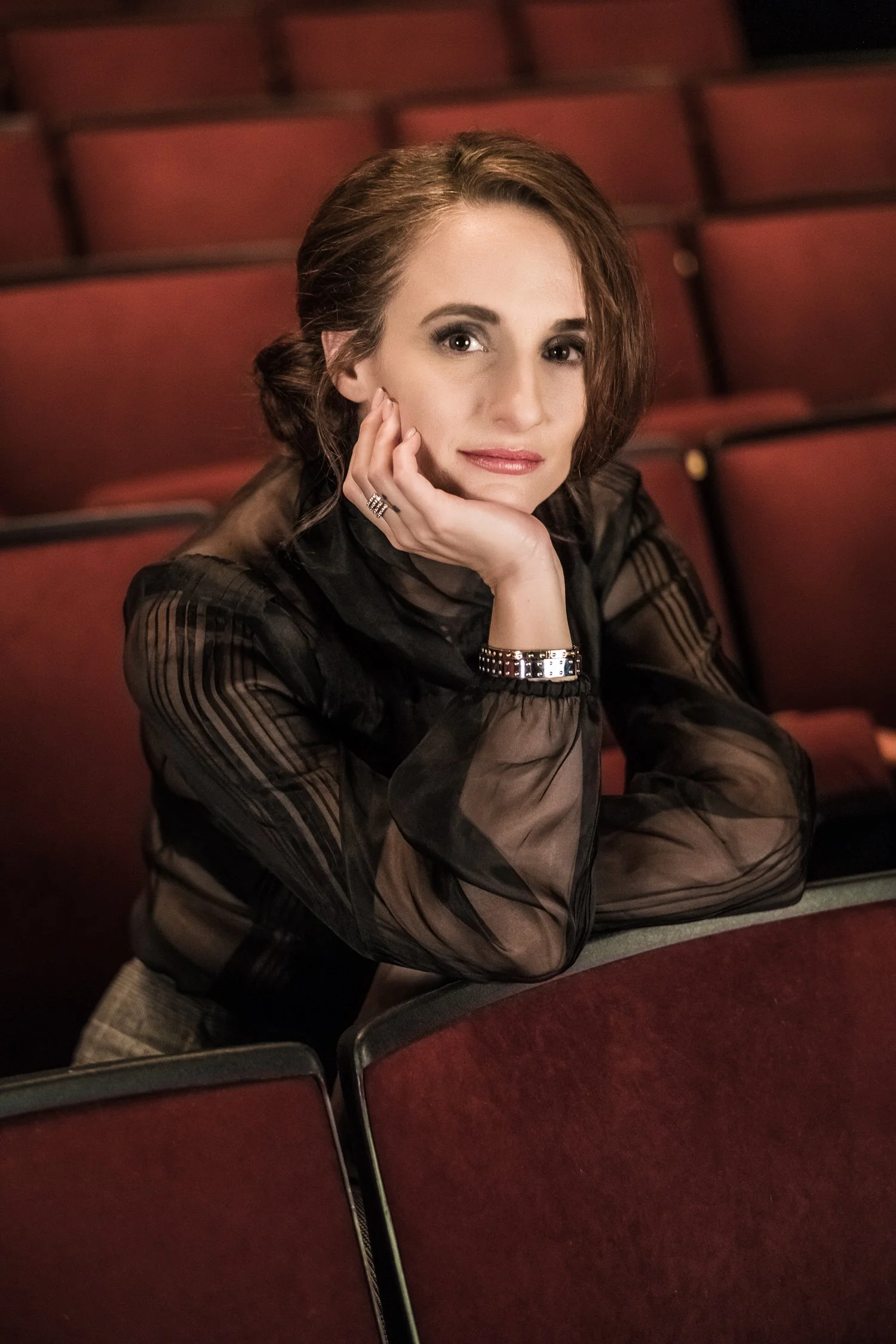 Actor Tracey Chimo with medium-length brown hair, sitting at a red theater or auditorium, resting her chin on her hand, looking at the camera.