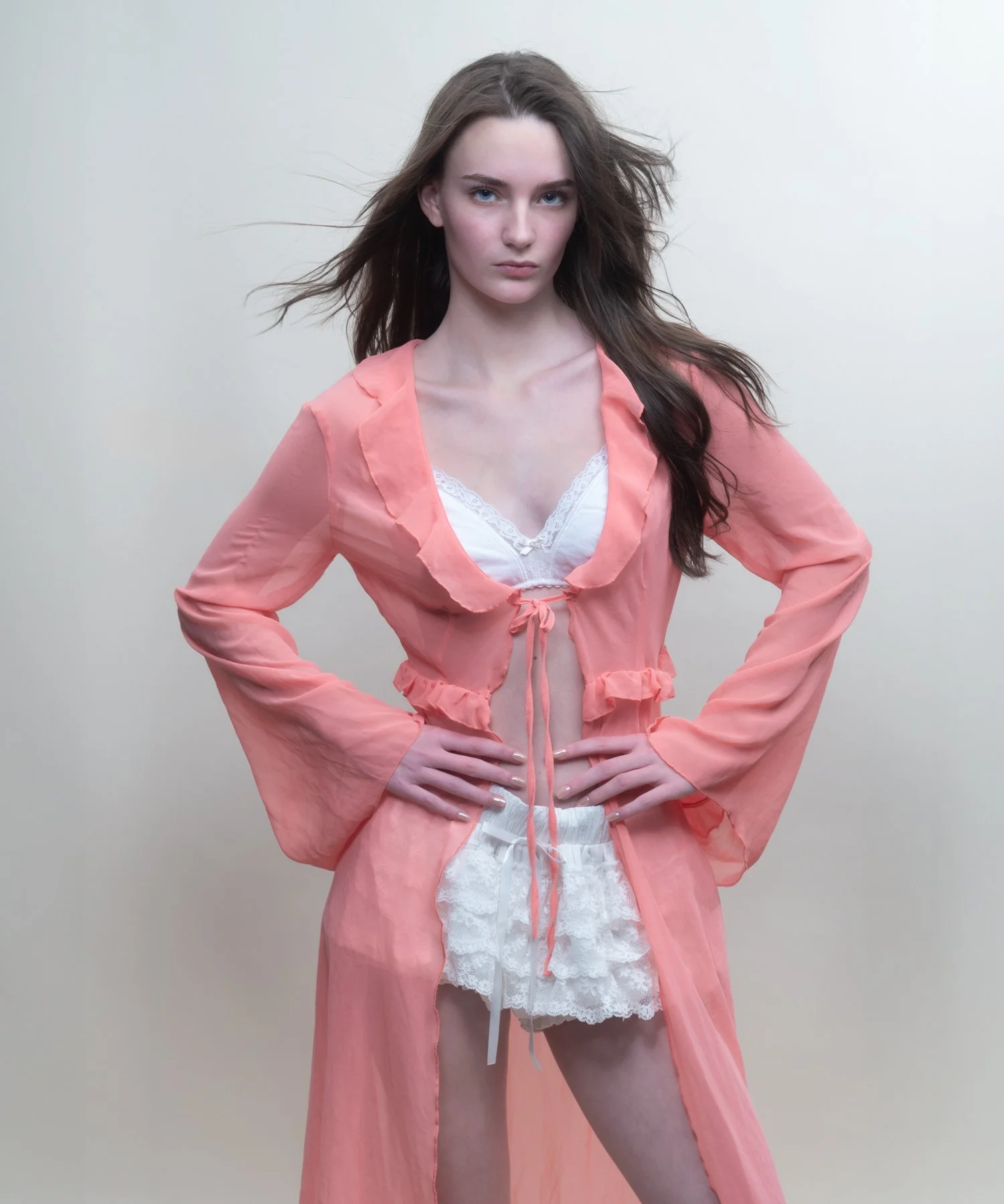 A young woman with long brown hair and blue eyes standing against a plain light-colored background, wearing a sheer pink robe over a white lace lingerie set, with her hands on her hips and an intense expression.