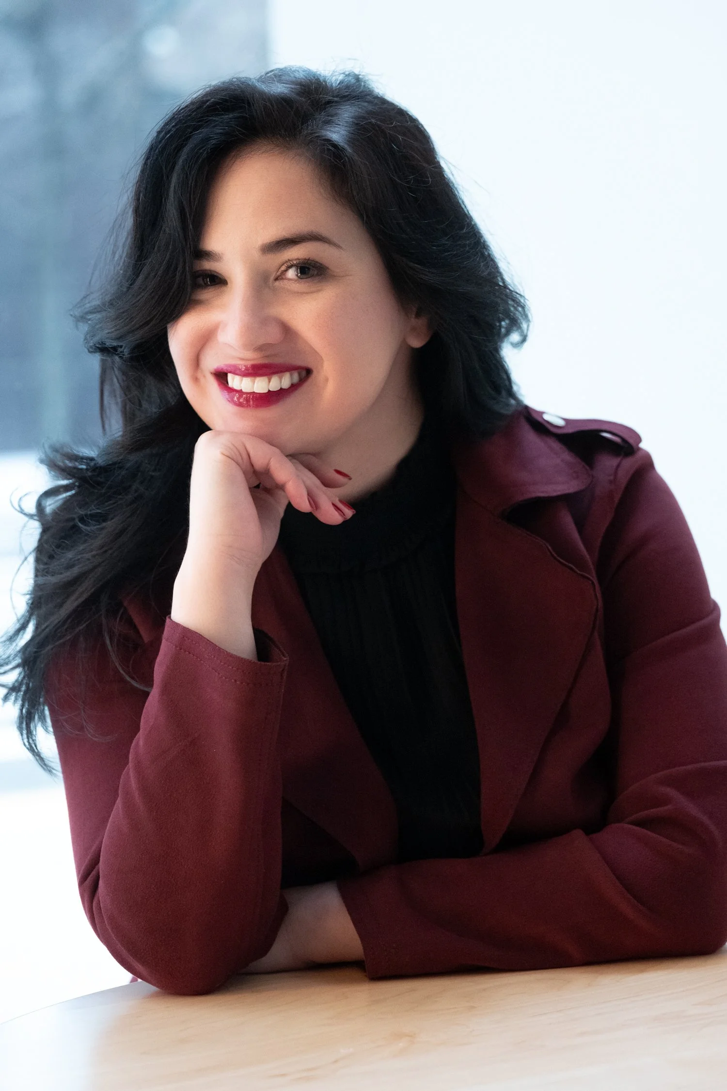 Smiling young woman with long, wavy black hair and red lips. She’s wearing a burgundy color jacket with her hand under her chin, photographed beside a window in natural light.