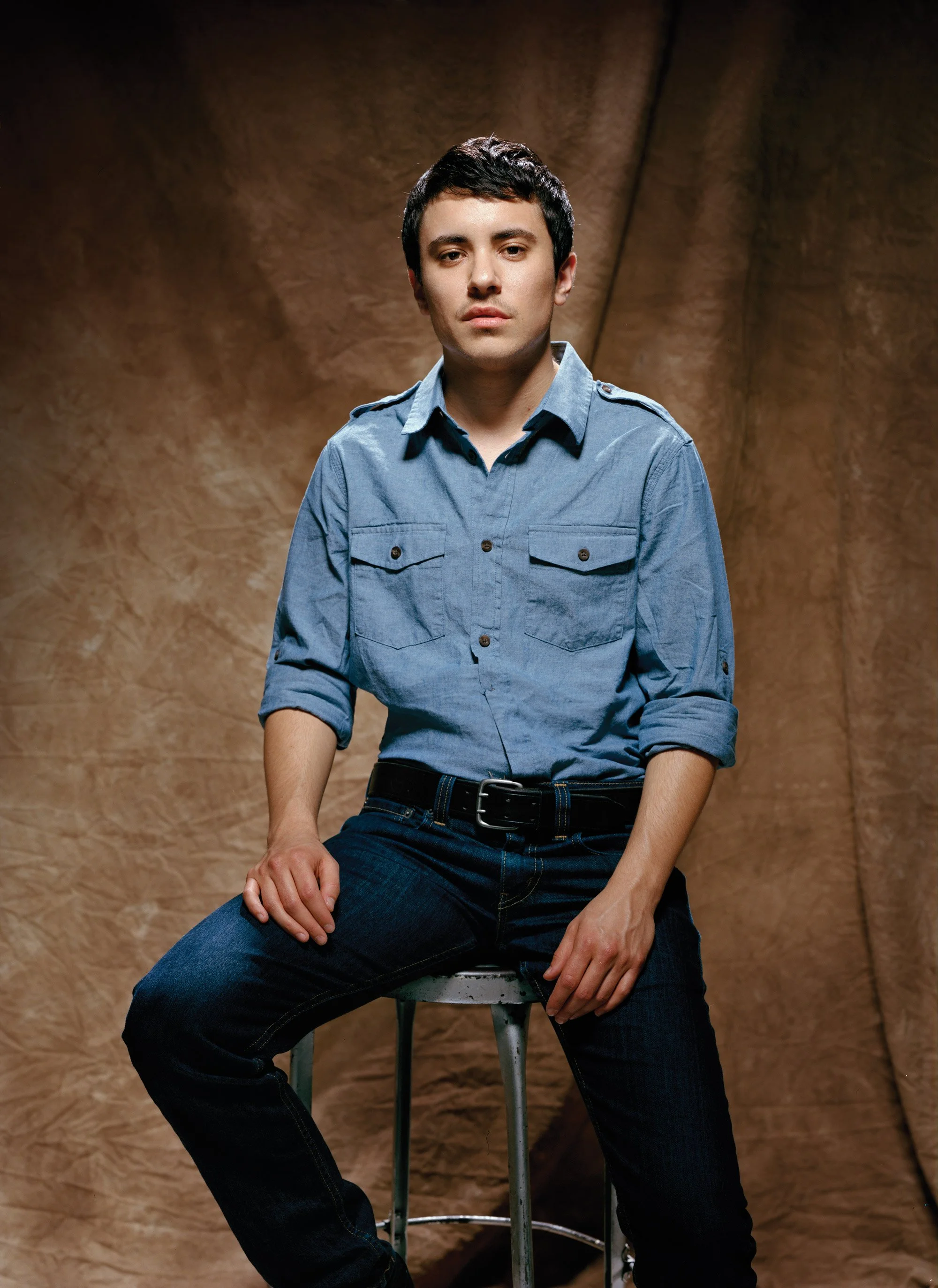 Young man with dark hair, wearing a blue denim shirt and jeans, sitting on a stool against a brown textured background.