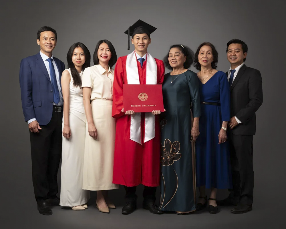 Group of seven people, including a graduate in red cap and gown holding a diploma, standing together in a studio portrait against a gray background, celebrating graduation from Boston University.