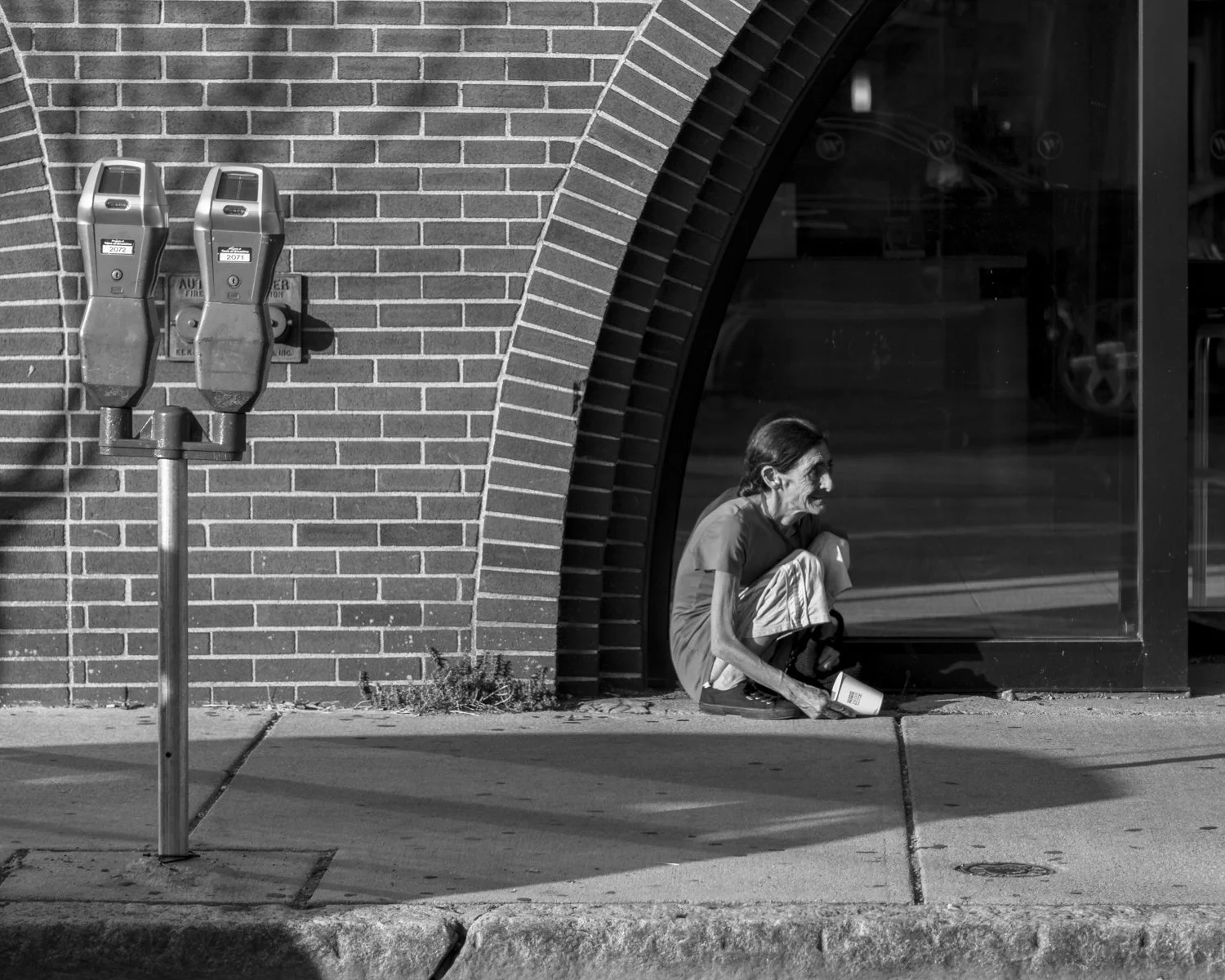 A homeless person sitting on the sidewalk outside a brick building, near large glass windows, with two parking meters nearby.