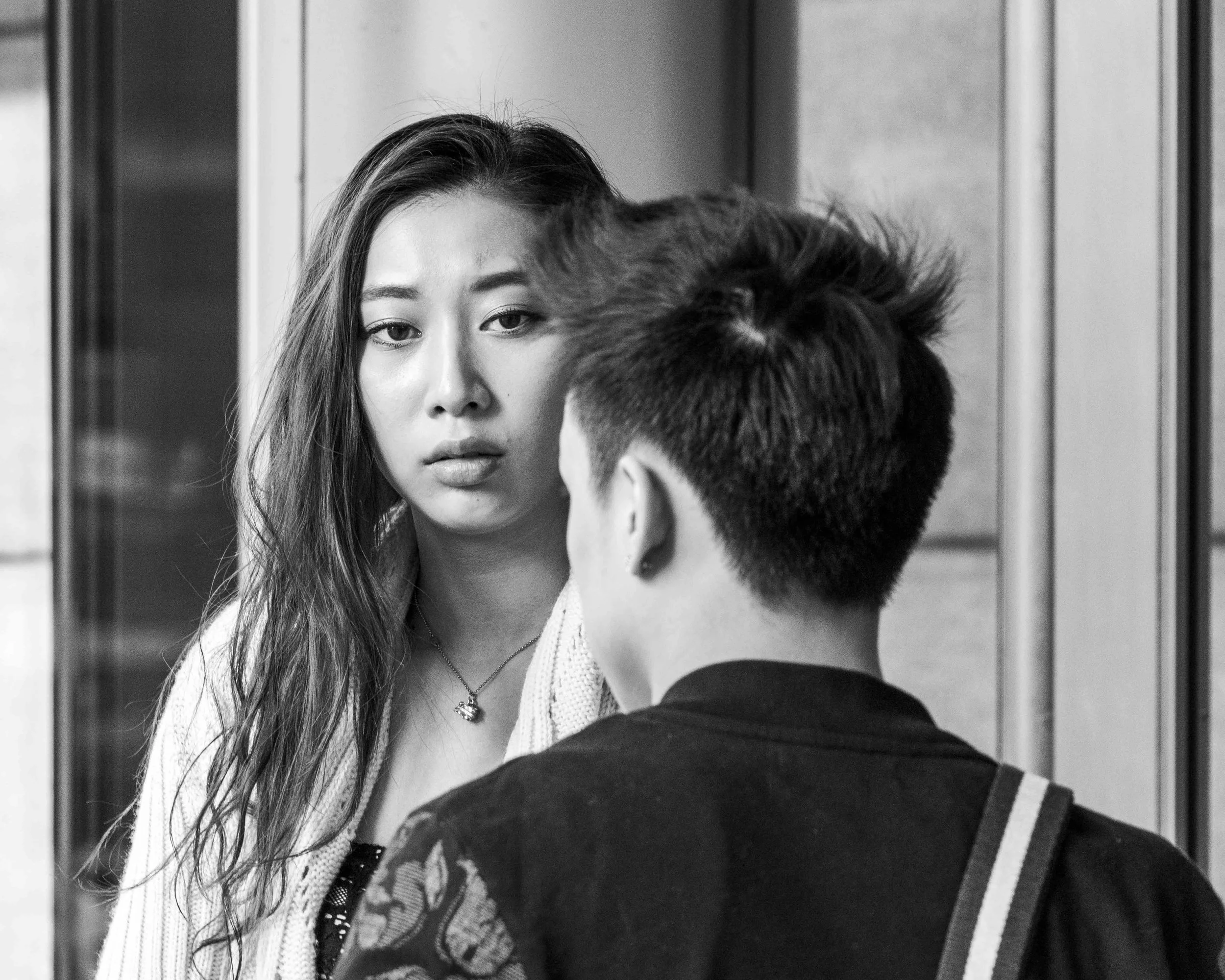 A black and white photograph of a young woman with long hair looking directly at the camera, with her face reflected in a mirror as she stands in a narrow corridor or elevator. The image captures her serious expression and the back of a person with s