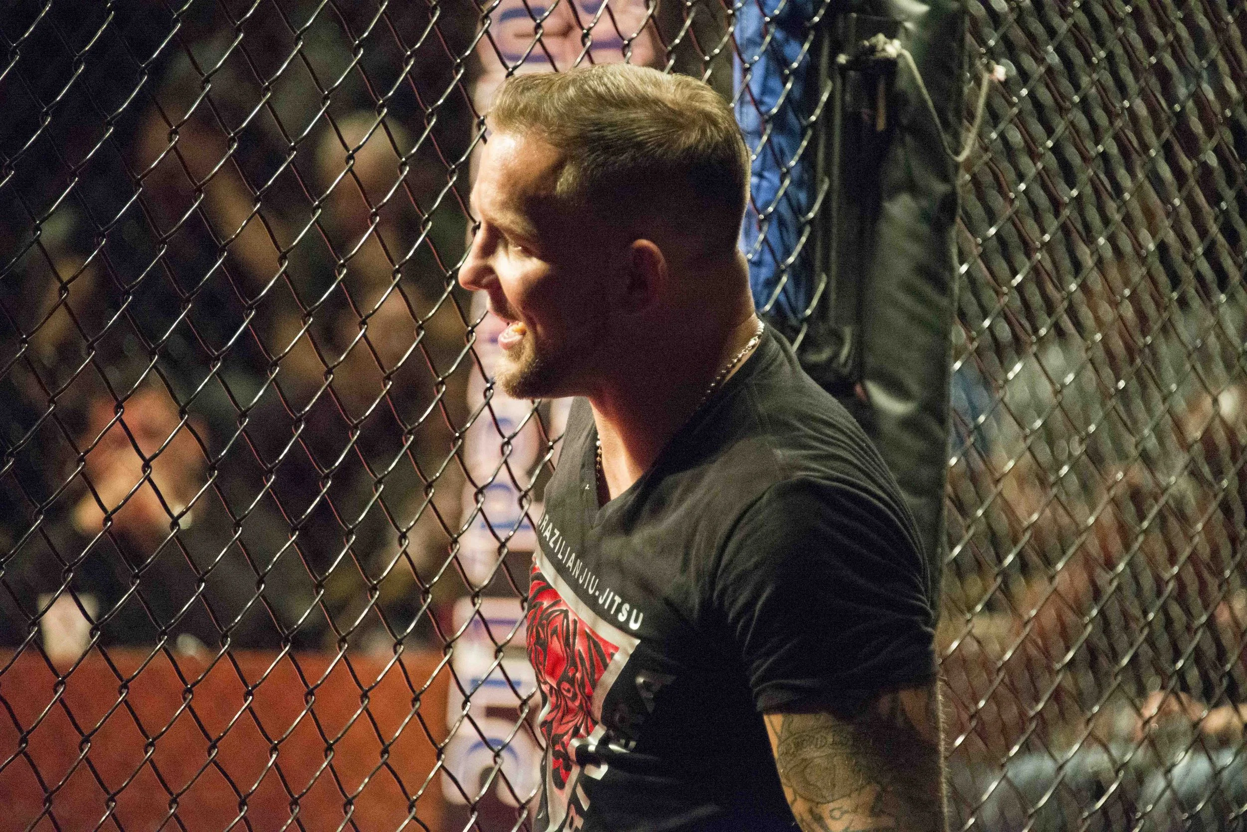 A man with short hair and tattoos on his arm, wearing a black t-shirt with red and white designs, sitting behind a chain-link fence at an MMA or boxing event, smiling and watching the action.