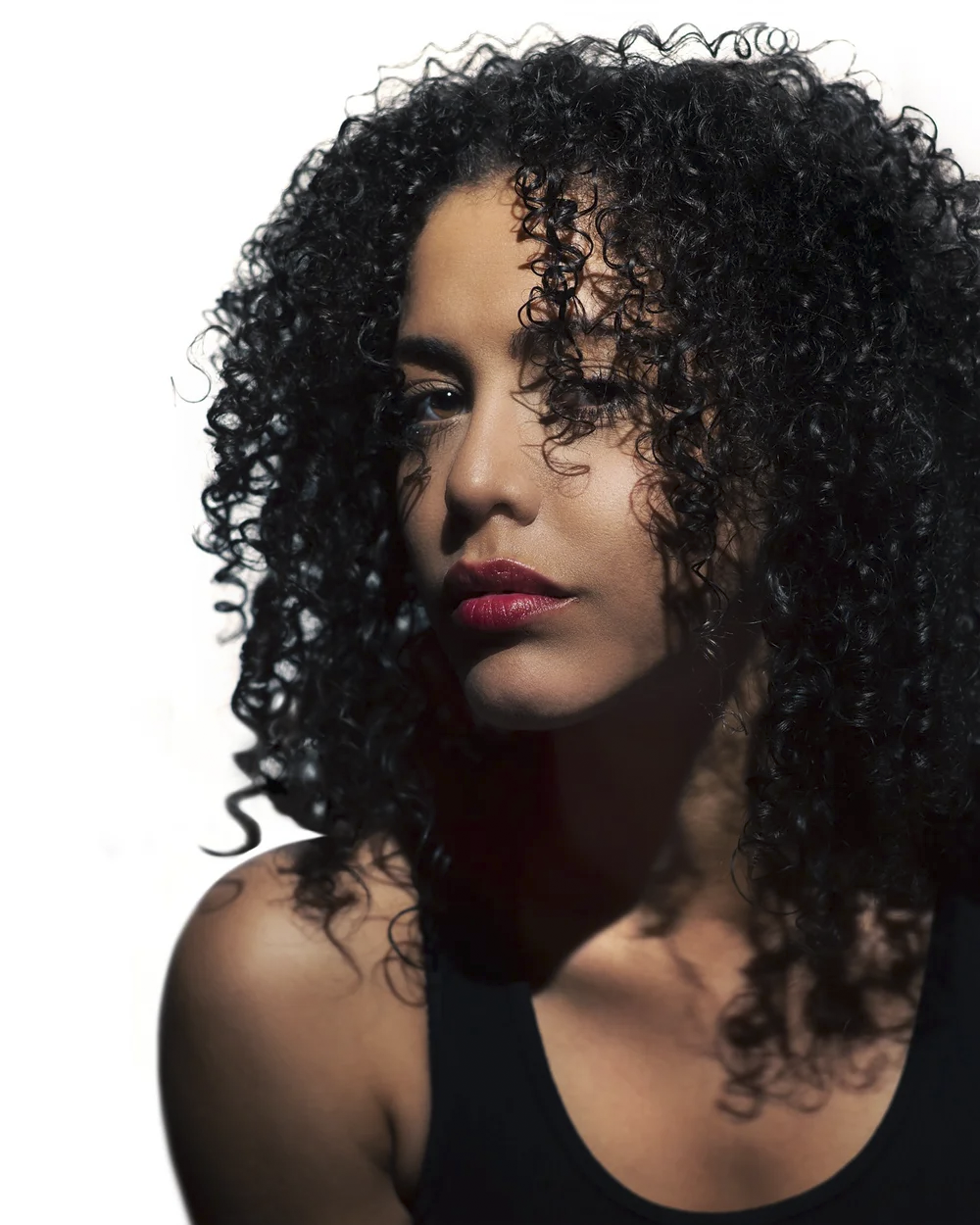 Close-up of a Latina woman with curly black hair, wearing a black sleeveless top, with a neutral white background.