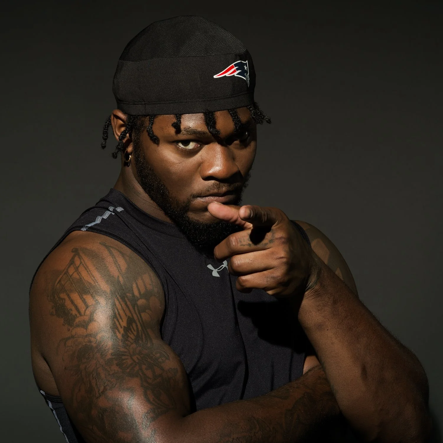 A muscular man with tattoos on his arm, wearing a black sleeveless shirt and a black cap with a sports team logo, pointing directly at the camera with an intense expression.