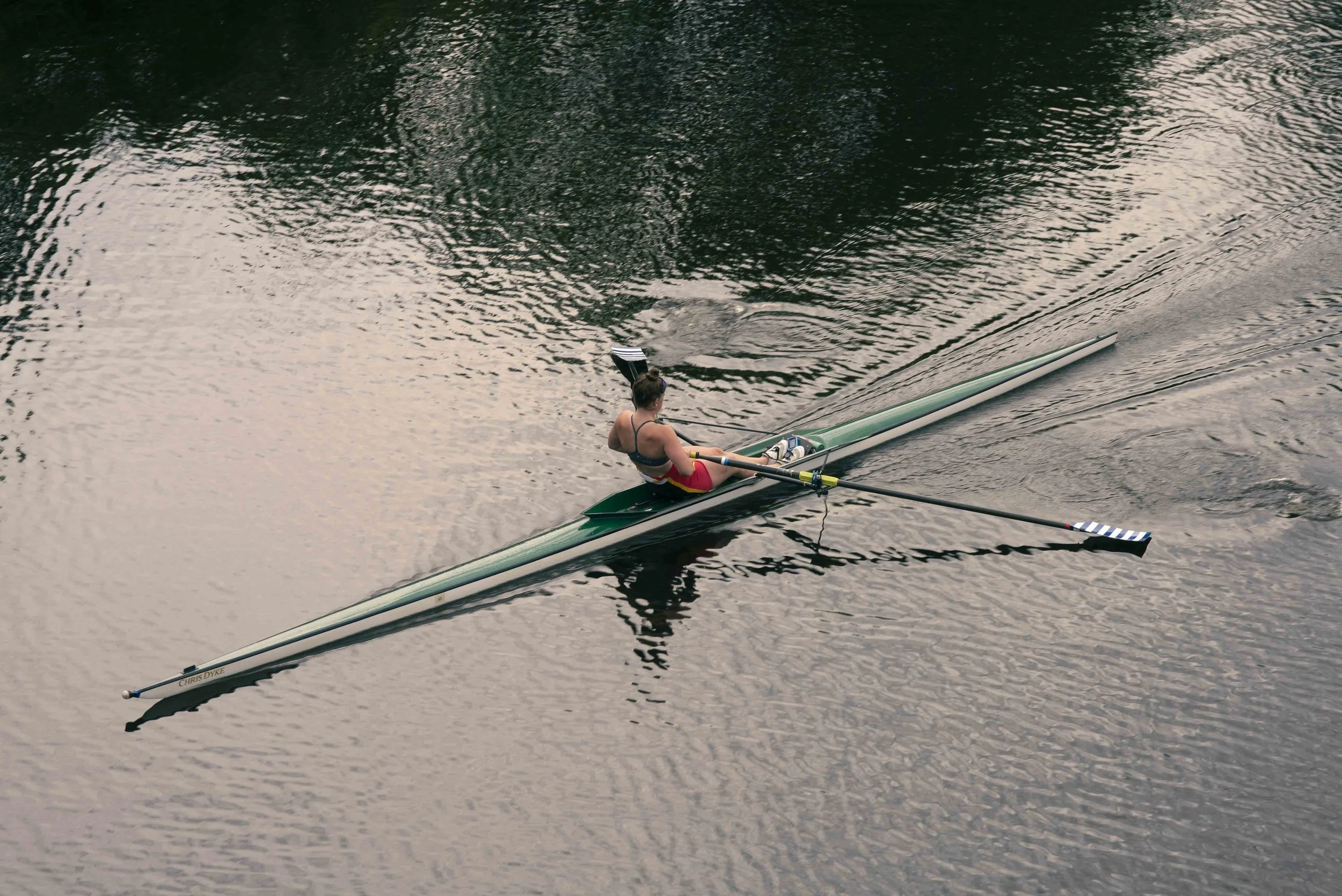 A person in a single scull rowing boat on a calm body of water, seen from above. The rower is wearing a black sports bra and red shorts, and is using two oars to propel the boat.