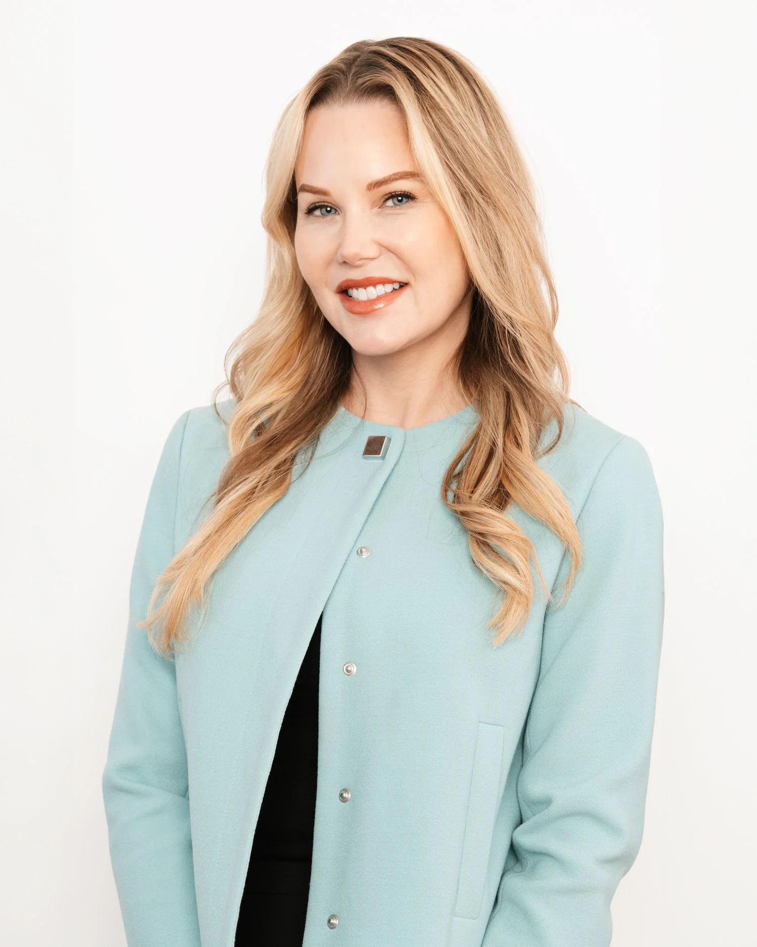 Upper body headshot of young, blond professional woman with orange lipstick in a seafoam aqua Mao-style jacket over a white background.
