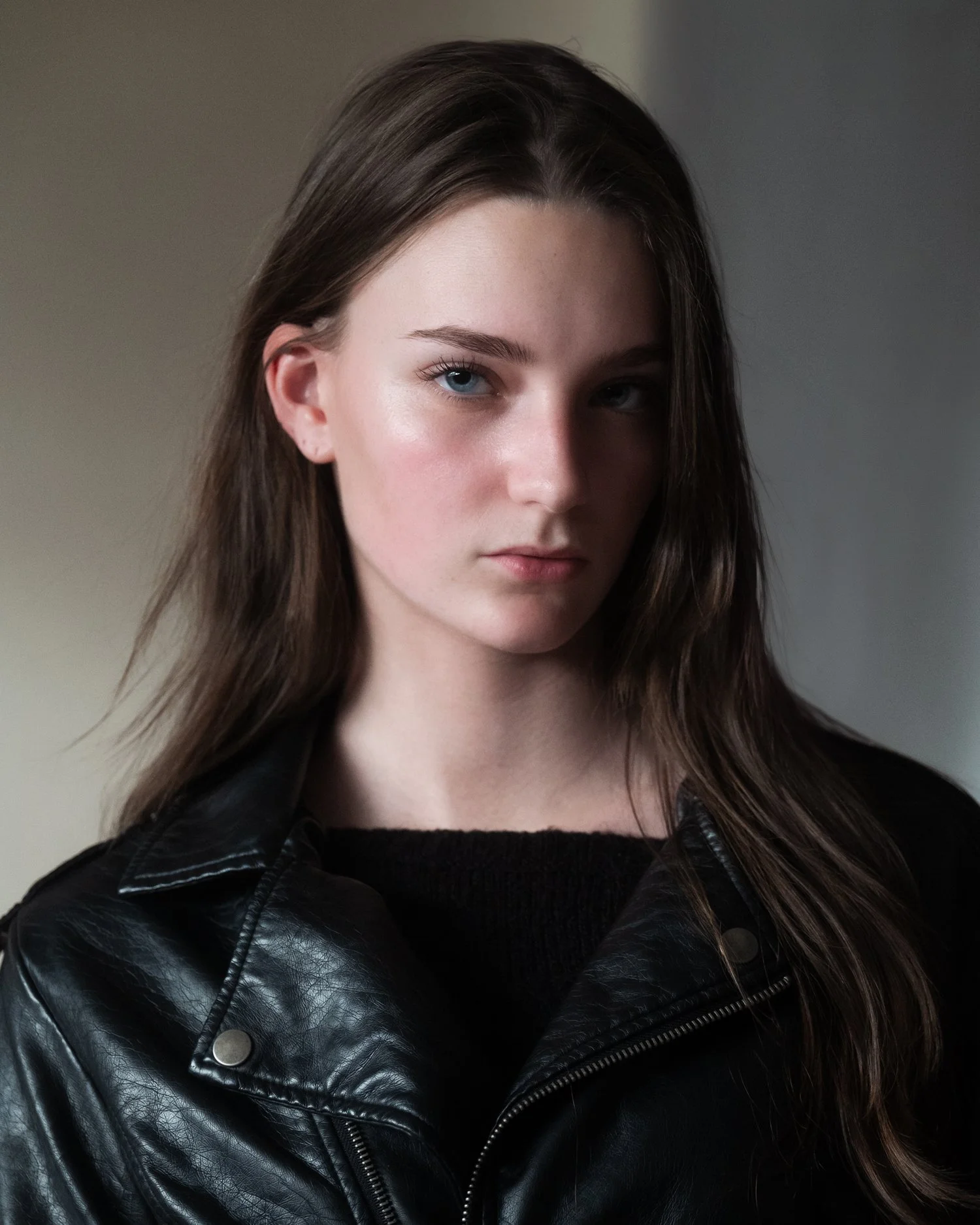 A young woman with long brown hair and blue eyes, wearing a black leather jacket, looking at the camera with a neutral expression.