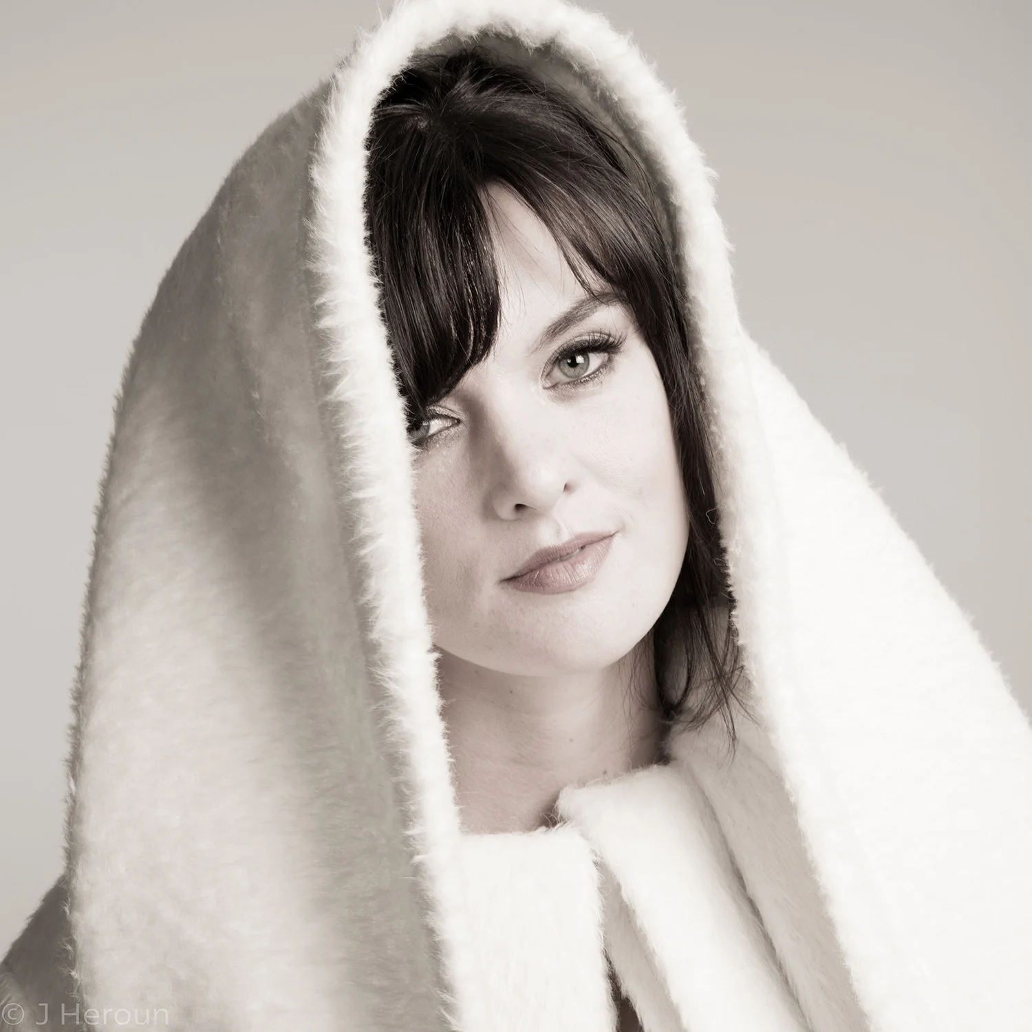 A woman with dark hair and light skin, wearing a white, fuzzy hooded garment, looking at the camera with a relaxed expression.