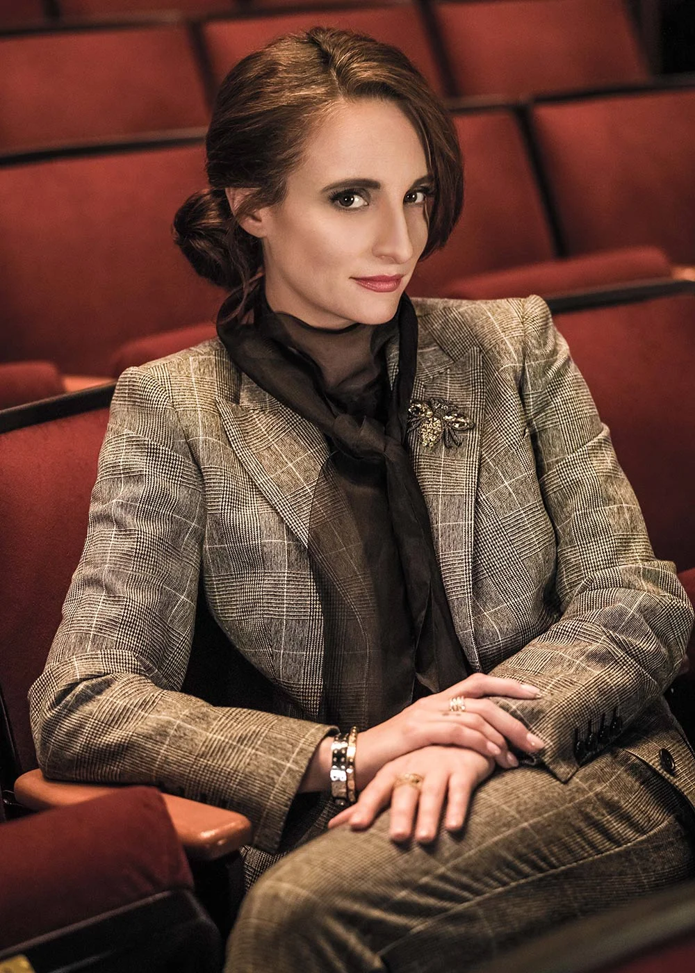 A woman with shoulder-length brown hair, wearing a plaid blazer and a black scarf, sitting in an auditorium with red seats.