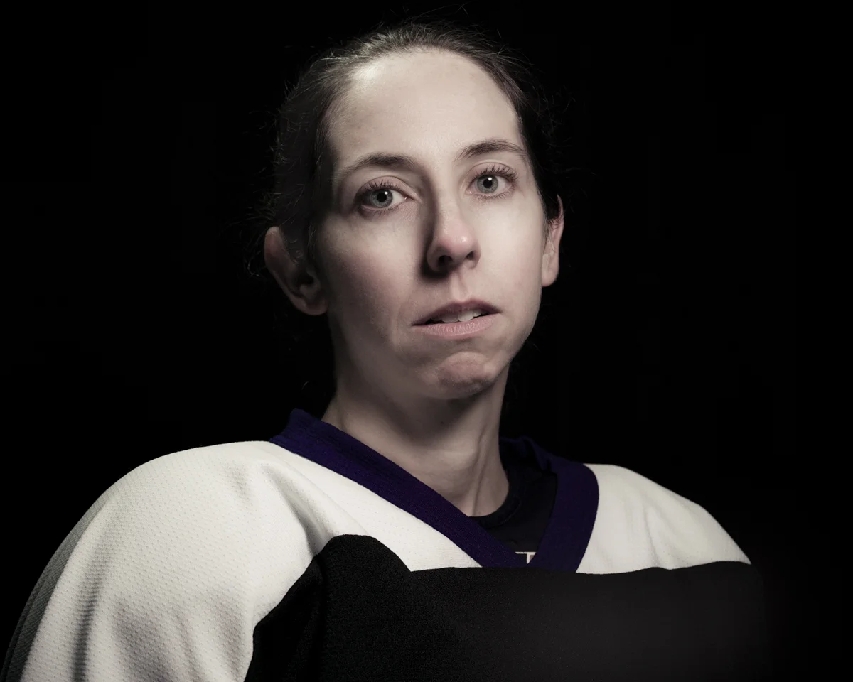 Portrait of a young woman with a serious expression, wearing a white sports jersey with purple and black accents, against a black background.