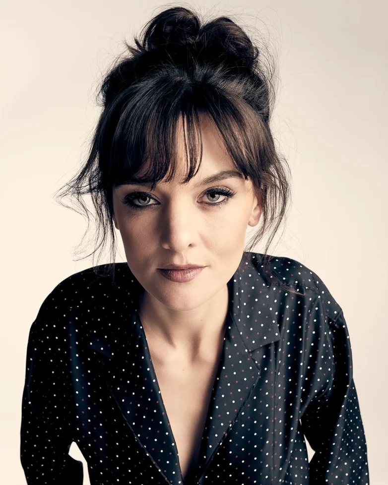 Portrait of a woman with dark hair styled in an updo, wearing a black blouse with white polka dots, looking directly at the camera.