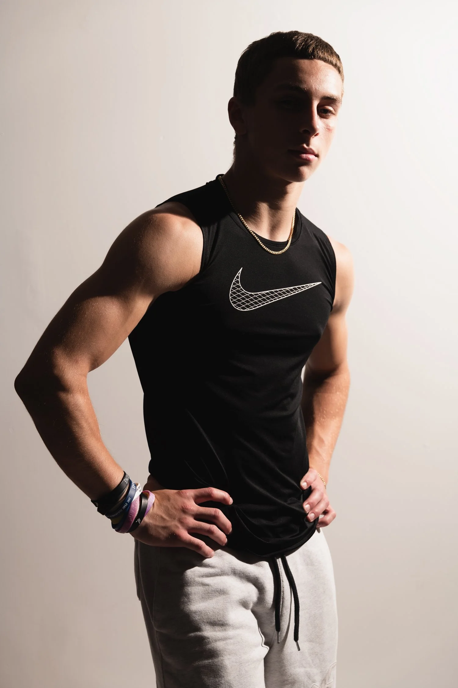 A young man in athletic wear, wearing a black Nike tank top and gray sweatpants, stands with his hands on his hips against a plain white background, accessorized with multiple bracelets and a necklace.