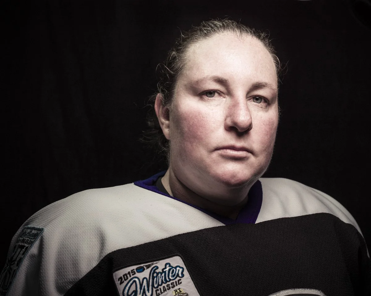 A woman in a hockey jersey with a serious expression, short hair pulled back, against a dark background.