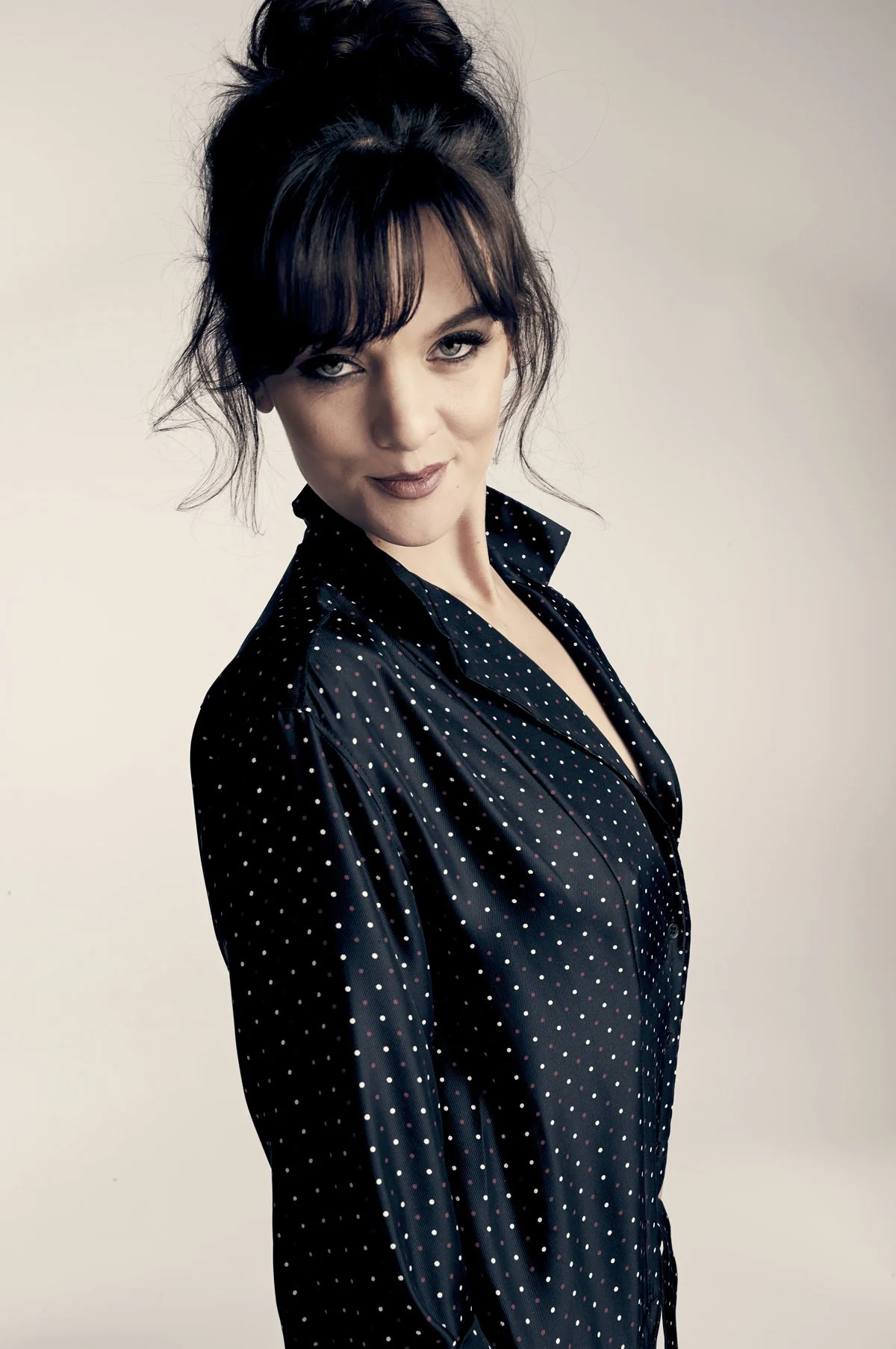 A woman with dark hair in a messy bun, wearing a black polka dot blouse, looking at the camera with a slight smile.