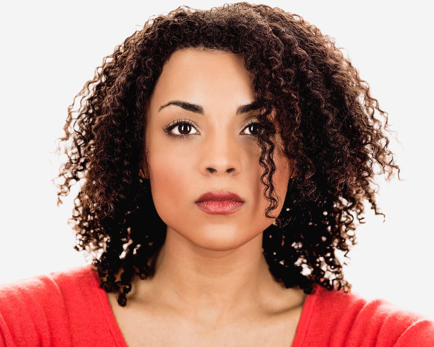 A Black woman with dark curly hair, wearing a red top, looking directly into the camera with a neutral expression against a white background.