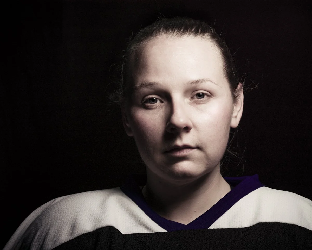 A young woman with light skin and brown hair in a ponytail poses against a black background, wearing a white and purple sports jersey.