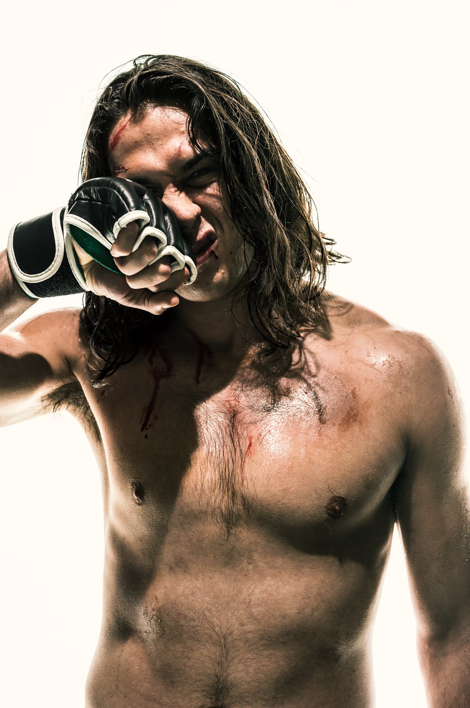 A shirtless male mixed martial artist with long dark hair, bruises and blood on his face, holding a glove near his face, appearing confident and fierce.