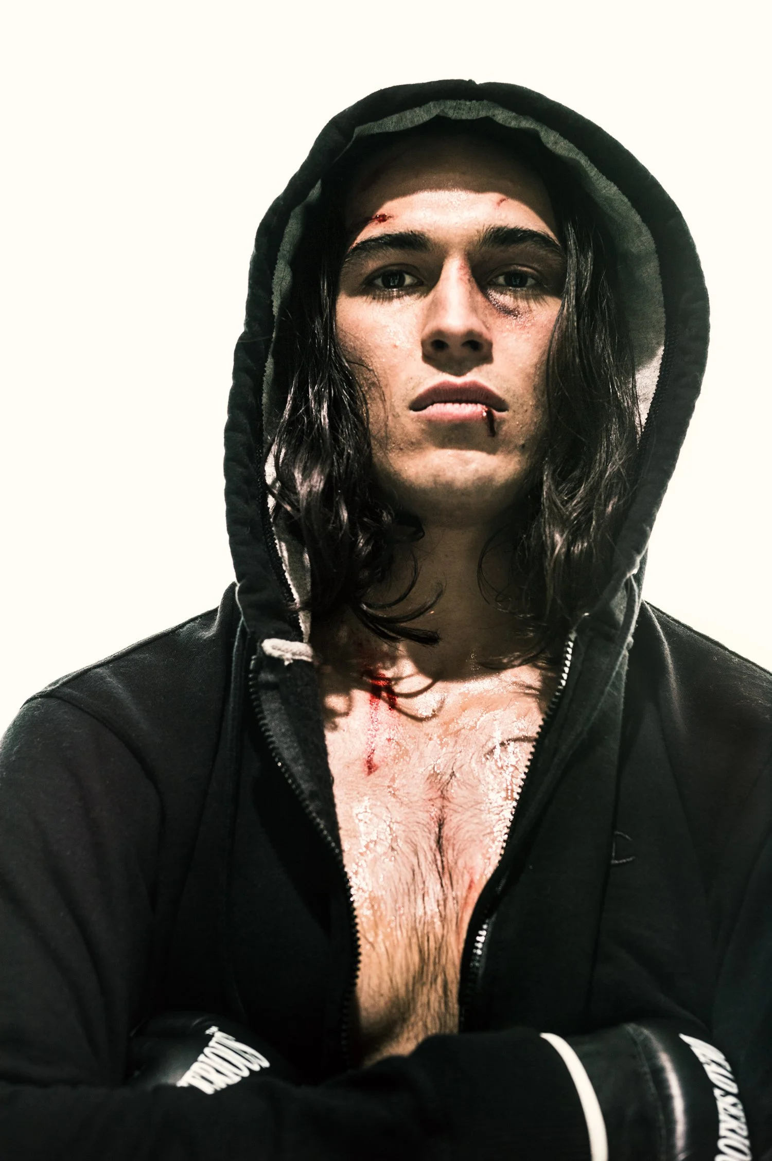 A young man with long, wet hair wearing a black hoodie with the hood up. He has visible injuries on his face, including a cut above his right eyebrow and a bruise around his right eye. His lips are slightly parted, and he has a cut on his lower lip. 