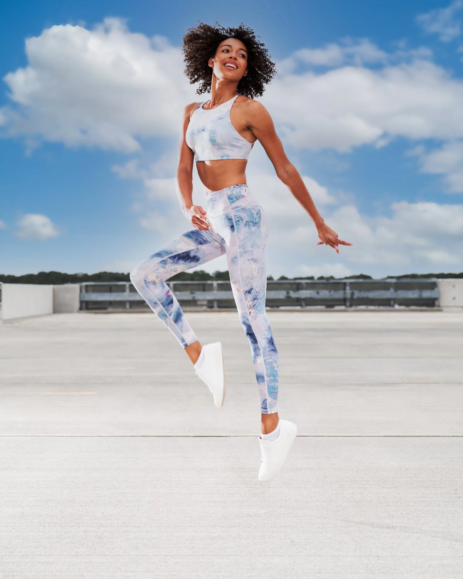A woman with curly hair wearing a sporty outfit and white sneakers, performing a dance or fitness move outdoors on a parking lot with a blue sky and clouds in the background.