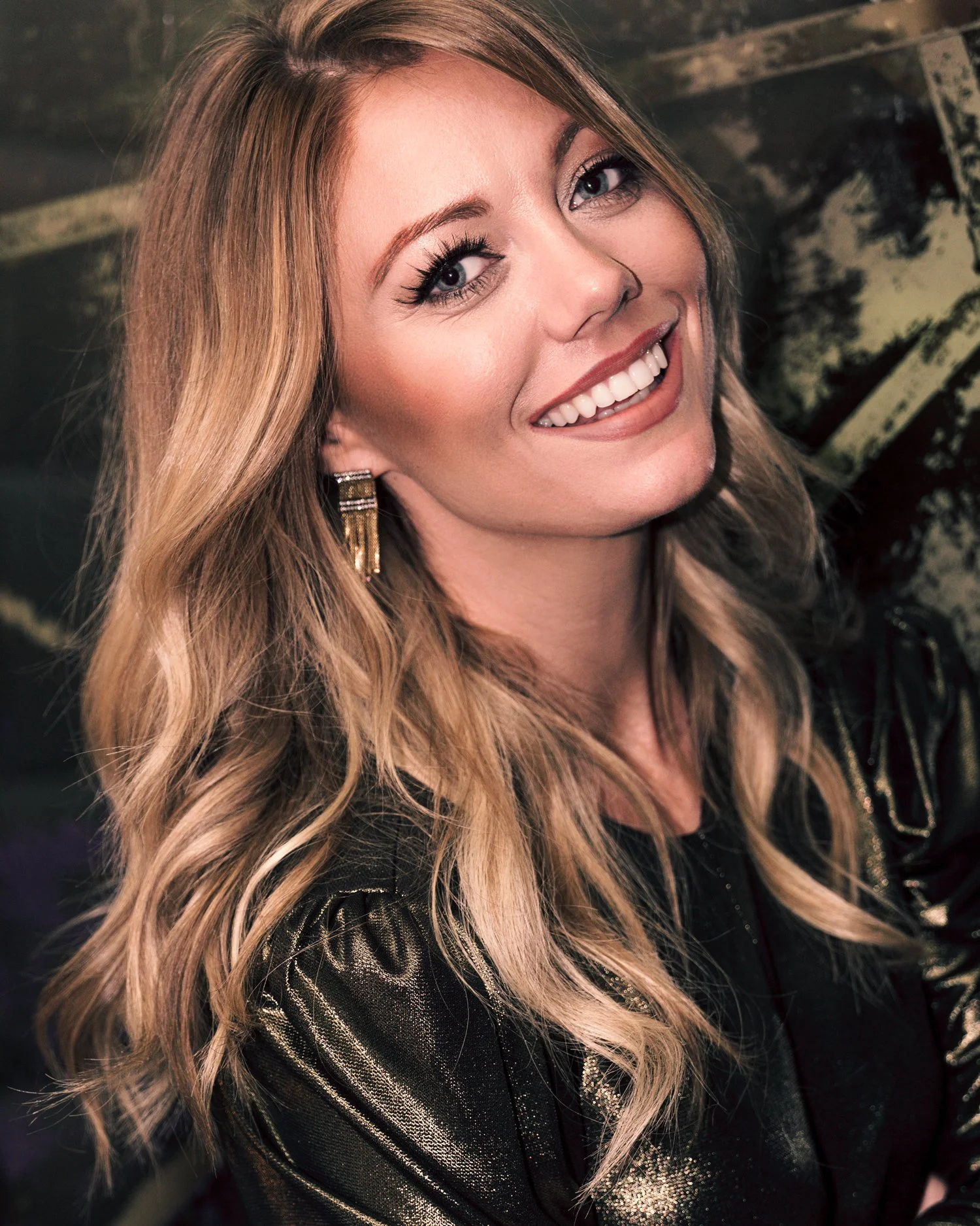 A smiling woman with long, wavy blonde hair, wearing makeup and gold earrings. She is dressed in a black outfit with metallic accents, standing in front of a dark, abstract background.