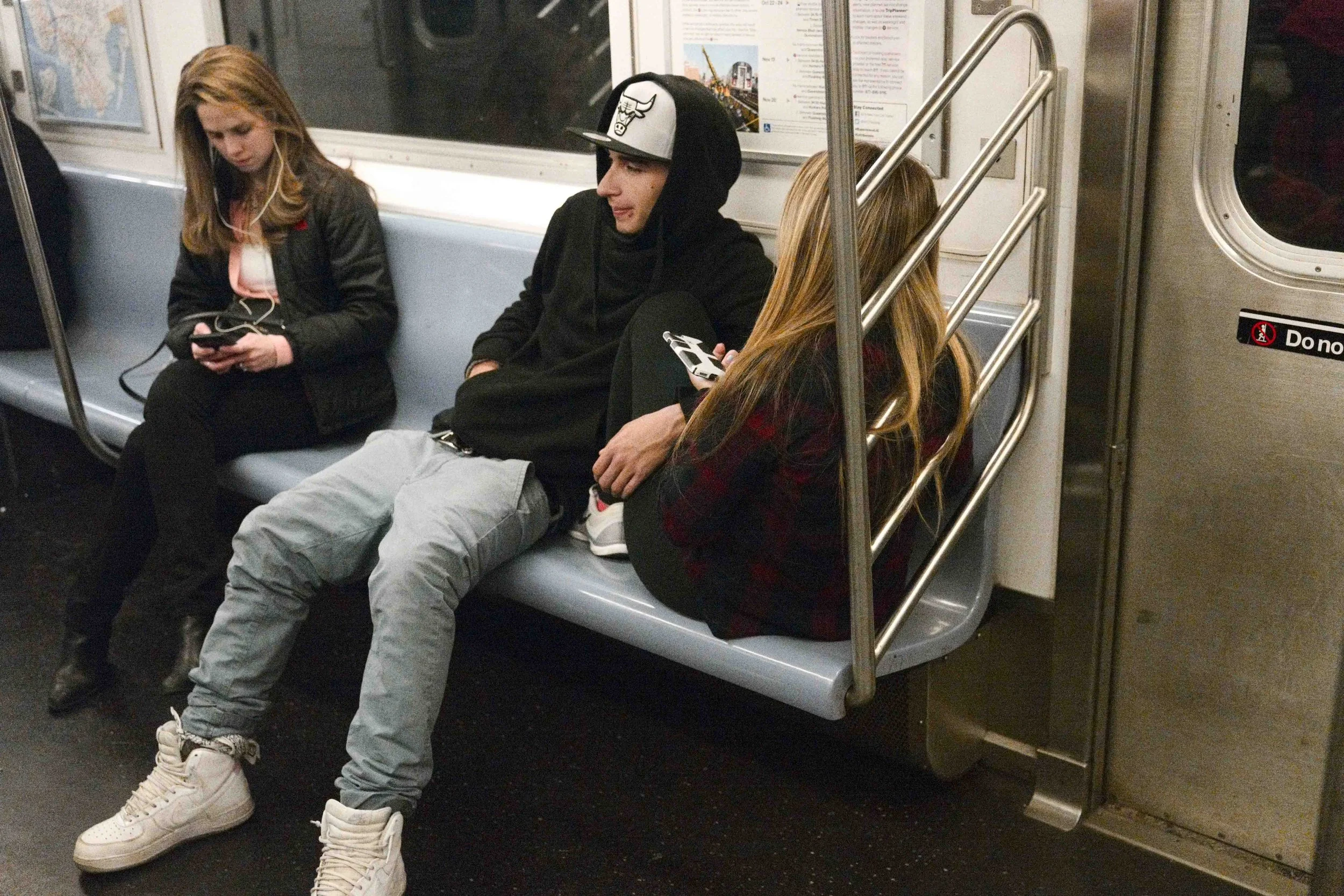 Three young people sitting on subway train seats, all looking at their phones, with one wearing a baseball cap and a hoodie, and two women with long hair, one with headphones.
