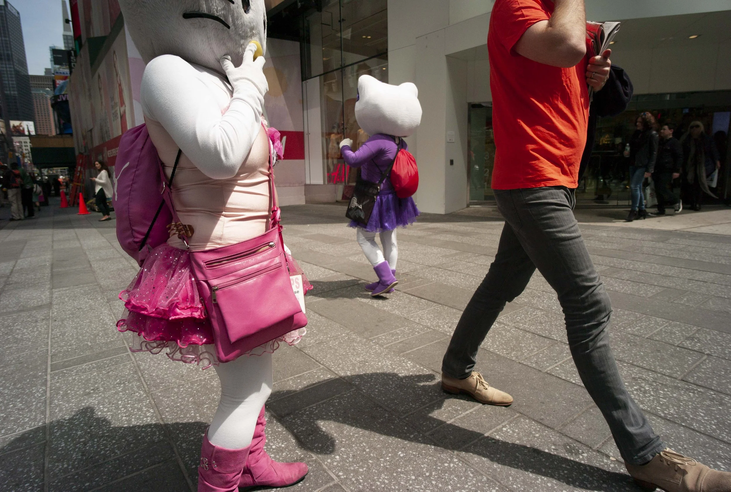 People walking on a city sidewalk, with a person dressed in a Hello Kitty mascot costume dressed in pink and white in the foreground.