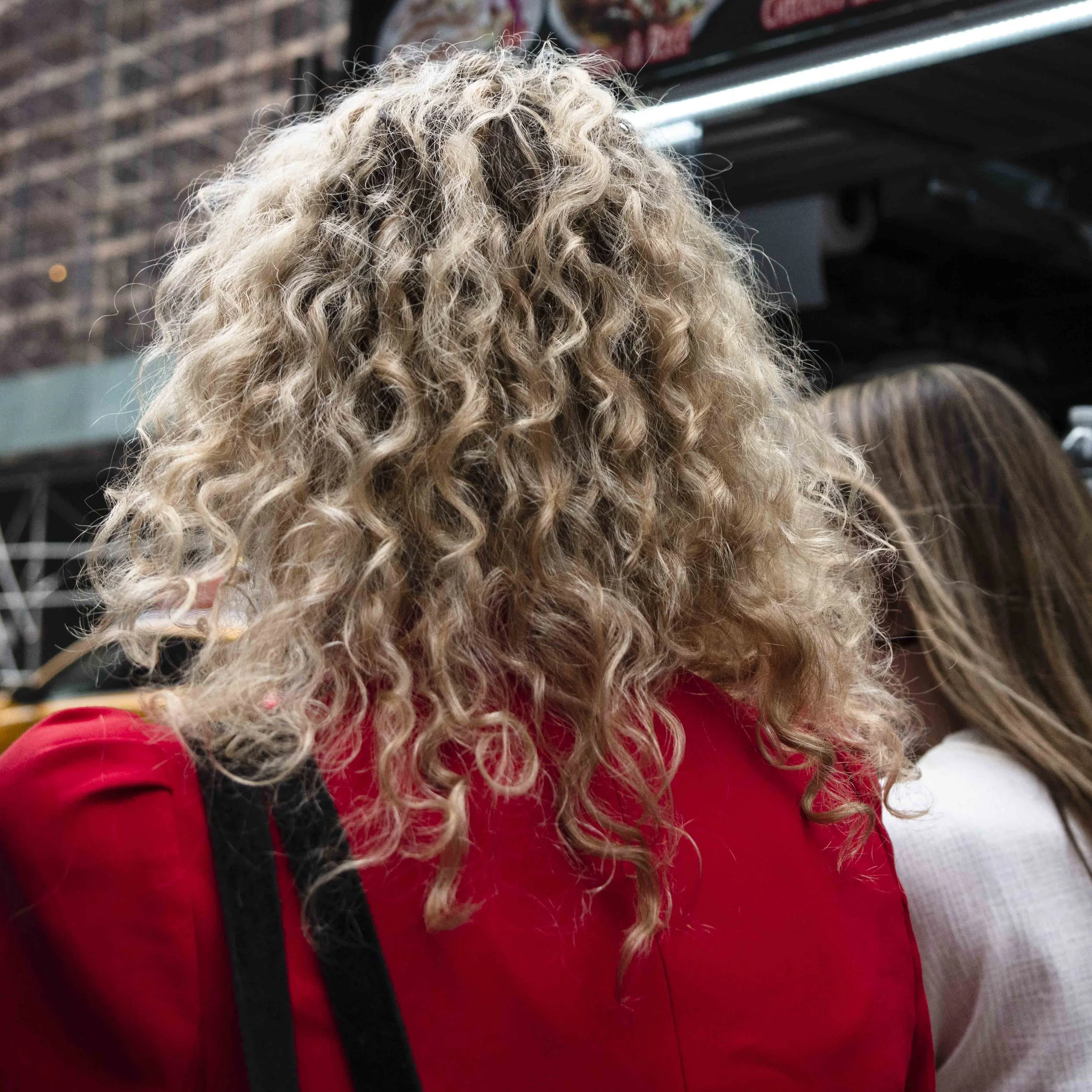 Back view of a person with long, curly blonde hair wearing a red jacket, in an indoor setting with shelves and objects in the background.