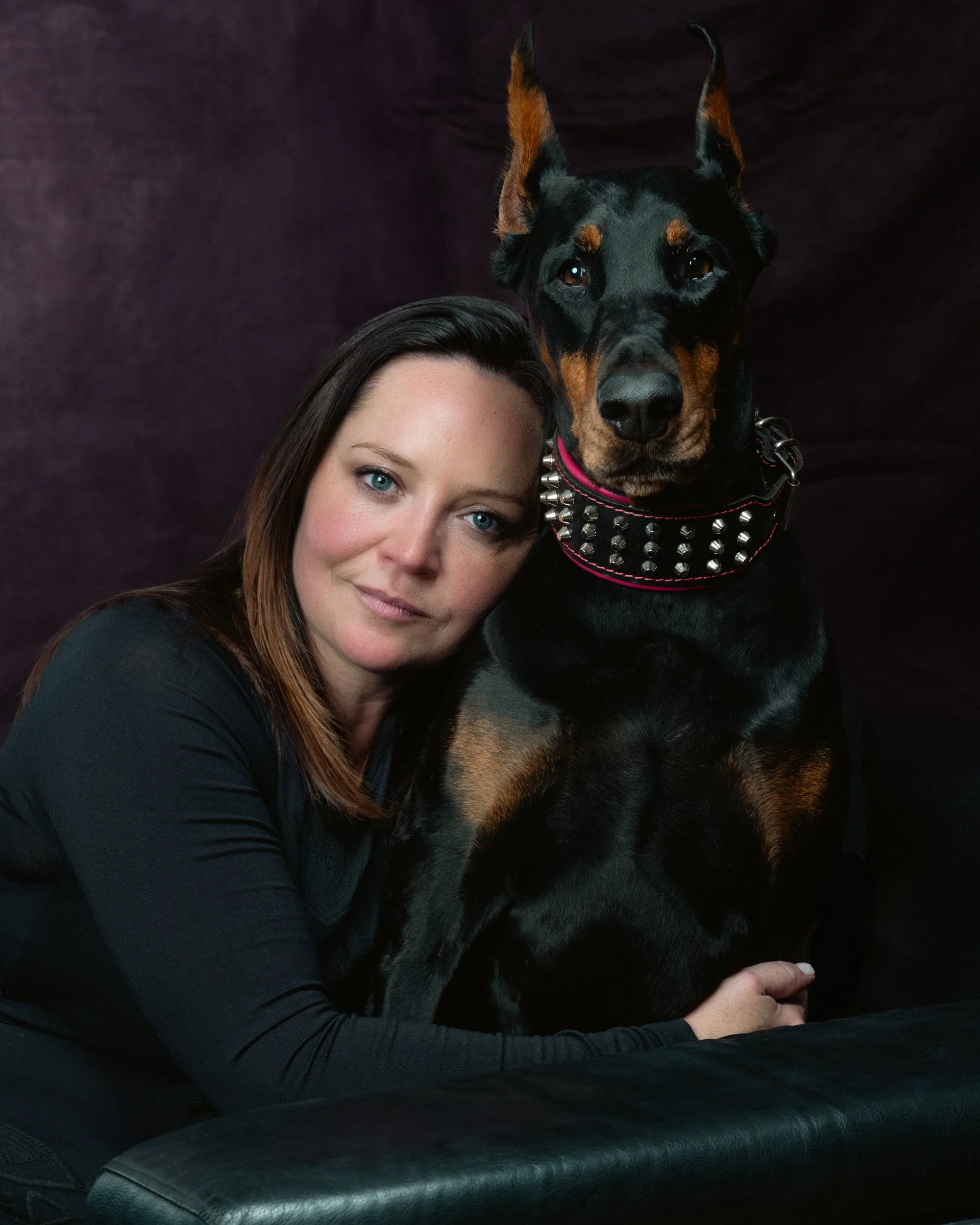 Formal studio portrait of a young woman with long, straight hair and black attire, posing with her black and tan Doberman canine against a dark burgundy-colored backdrop. The dog wears a large studded collar with red piping on the edges.