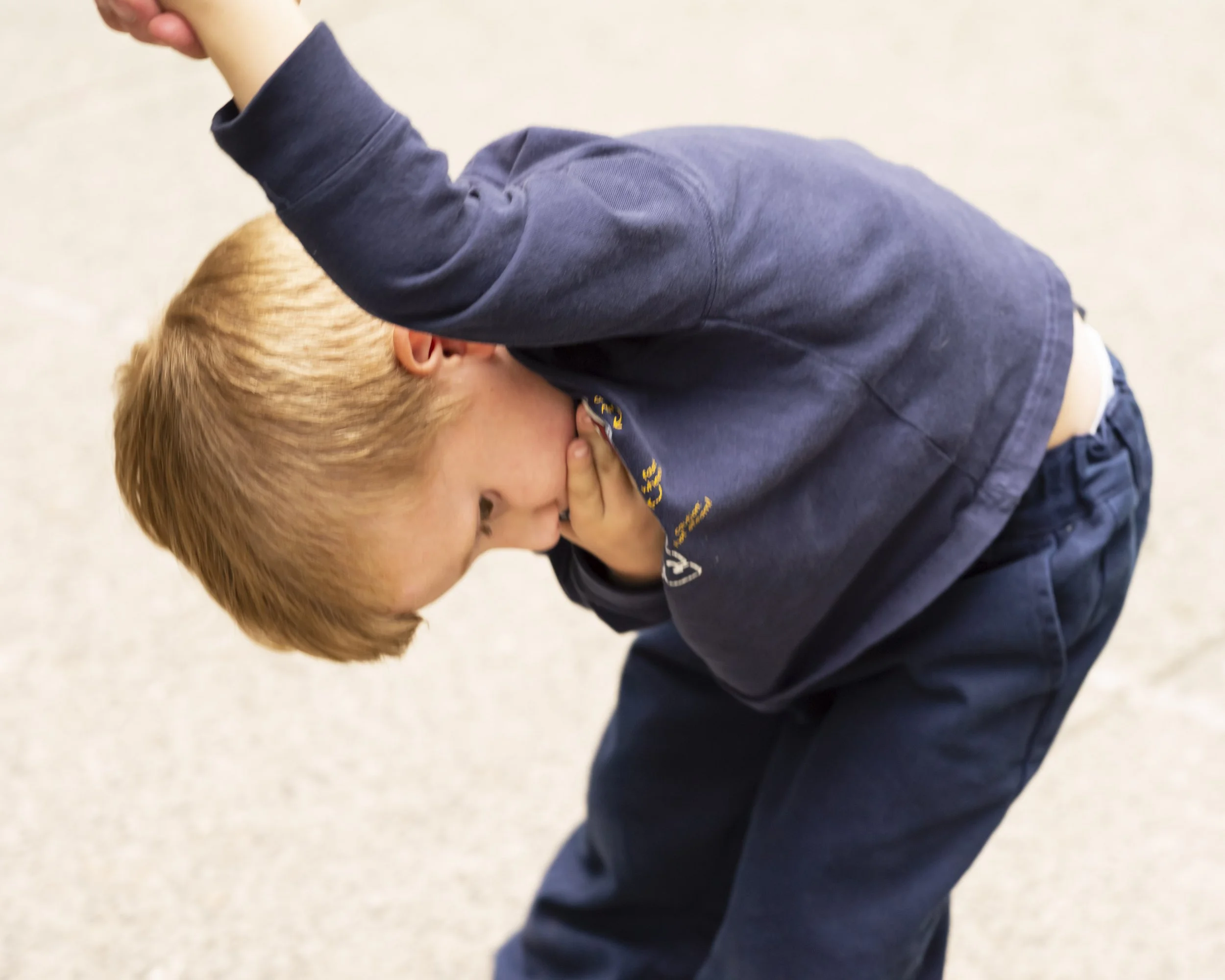 A young boy with blond hair wearing a dark jacket is crouching down and covering his face with his hand, standing on a light-colored surface.