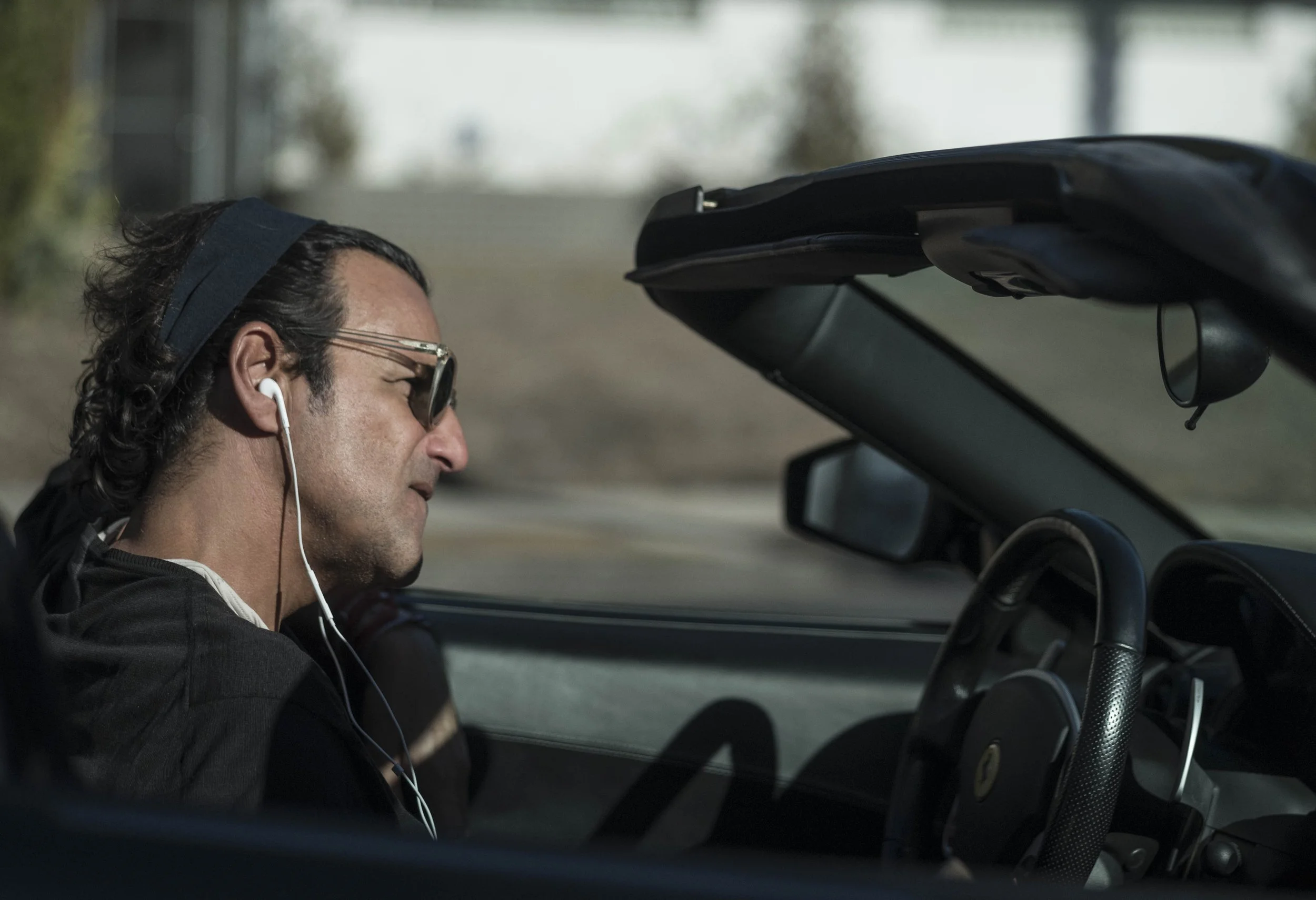 A man with dark hair, sunglasses, and earbuds sits in a convertible car, with the top down, at a stop light on a sunny day.