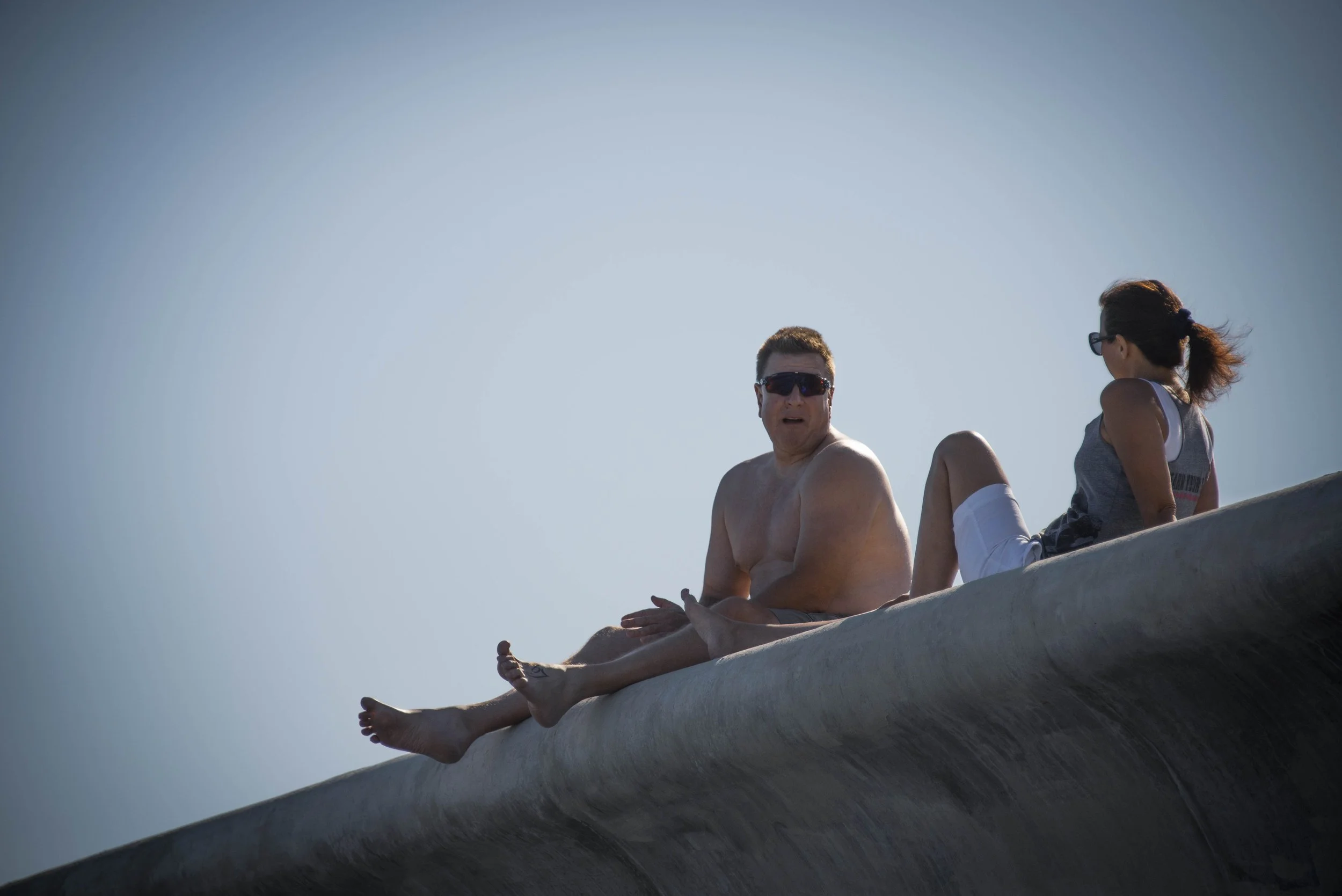 Two people sit on a large, rounded rock against a clear blue sky. One is a shirtless man wearing sunglasses and the other is a woman with glasses and a ponytail, dressed in casual summer clothing.