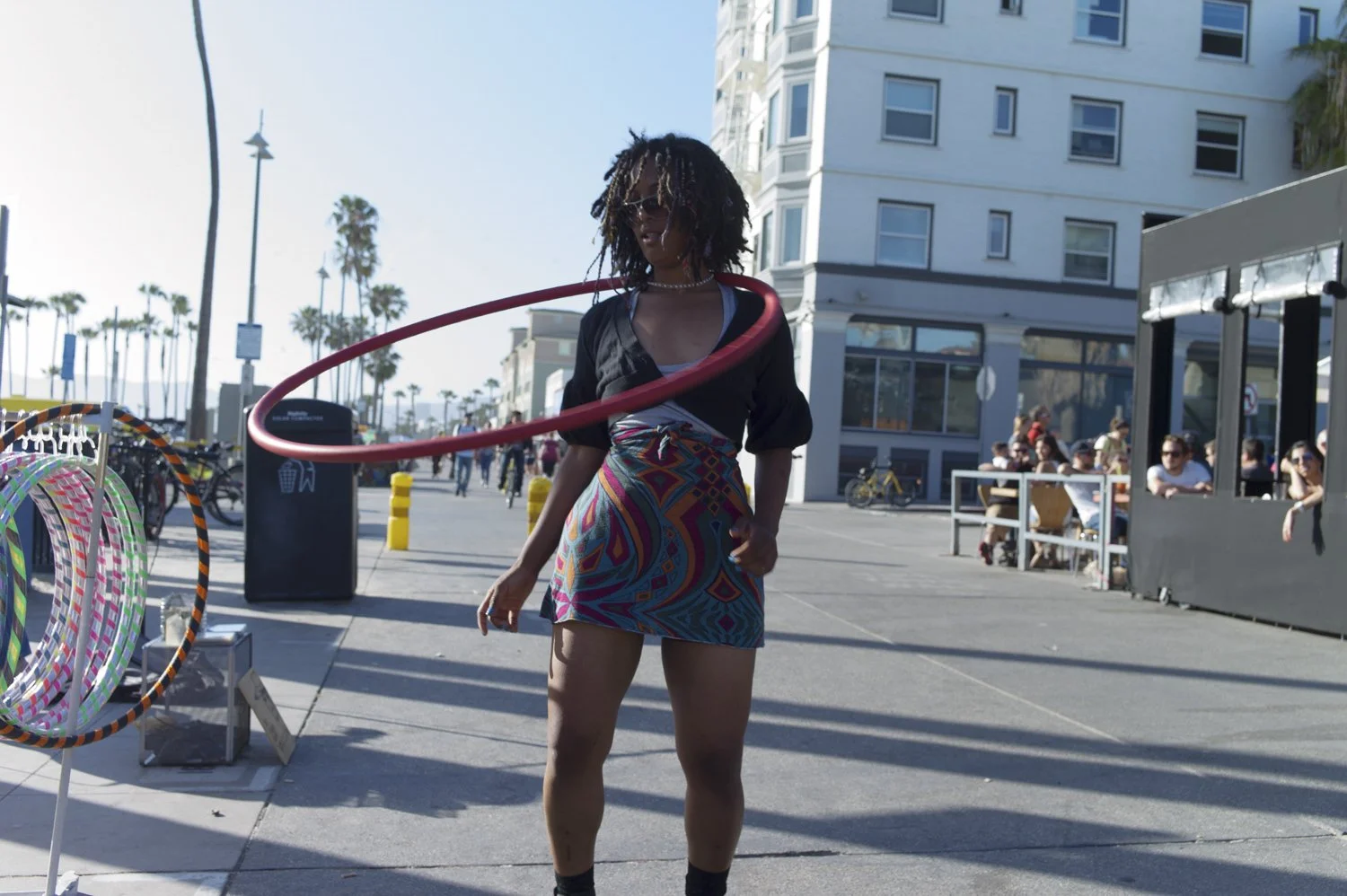 A woman with dark curly hair and sunglasses hula hooping on a sunny day near the beach, with a crowd of people sitting at outdoor tables and palm trees in the background.