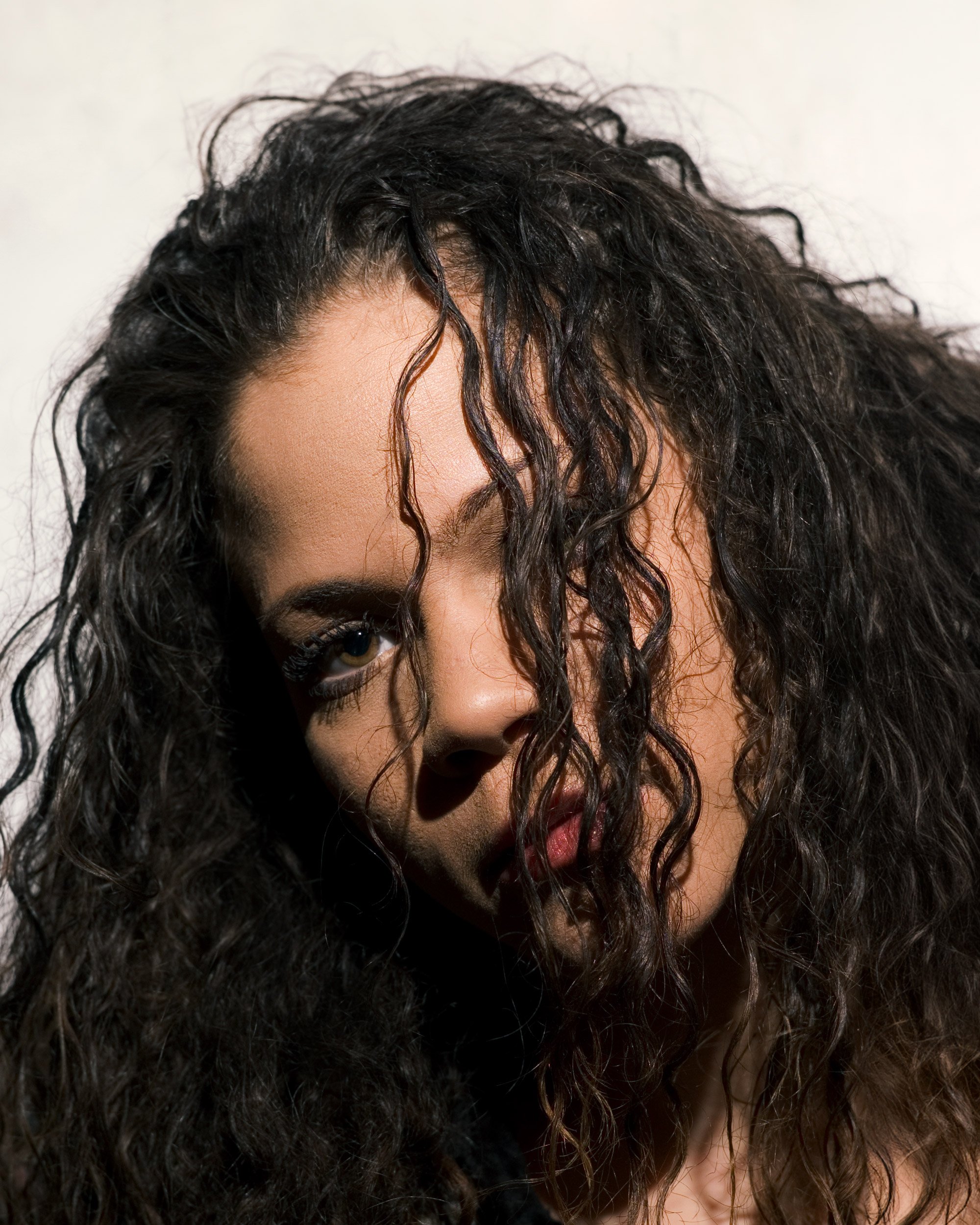 Close-up portrait of an intense young Latina with long, wavy hair obscuring most of her face.