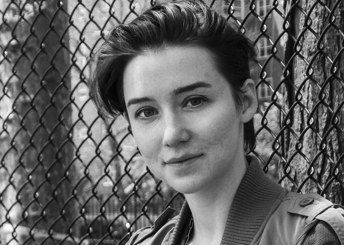 Black and white photo of a young woman with short hair smiling softly, standing in front of a chain-link fence with trees and buildings in the background.