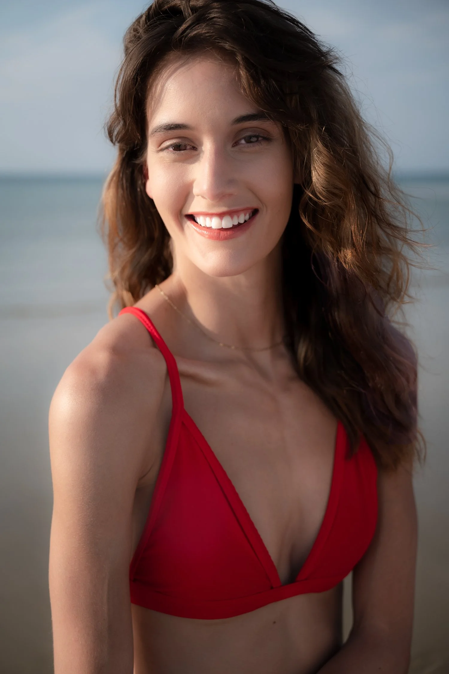A smiling young woman with brown, wavy hair, wearing a red bikini top, standing on a beach with a blurred ocean background.